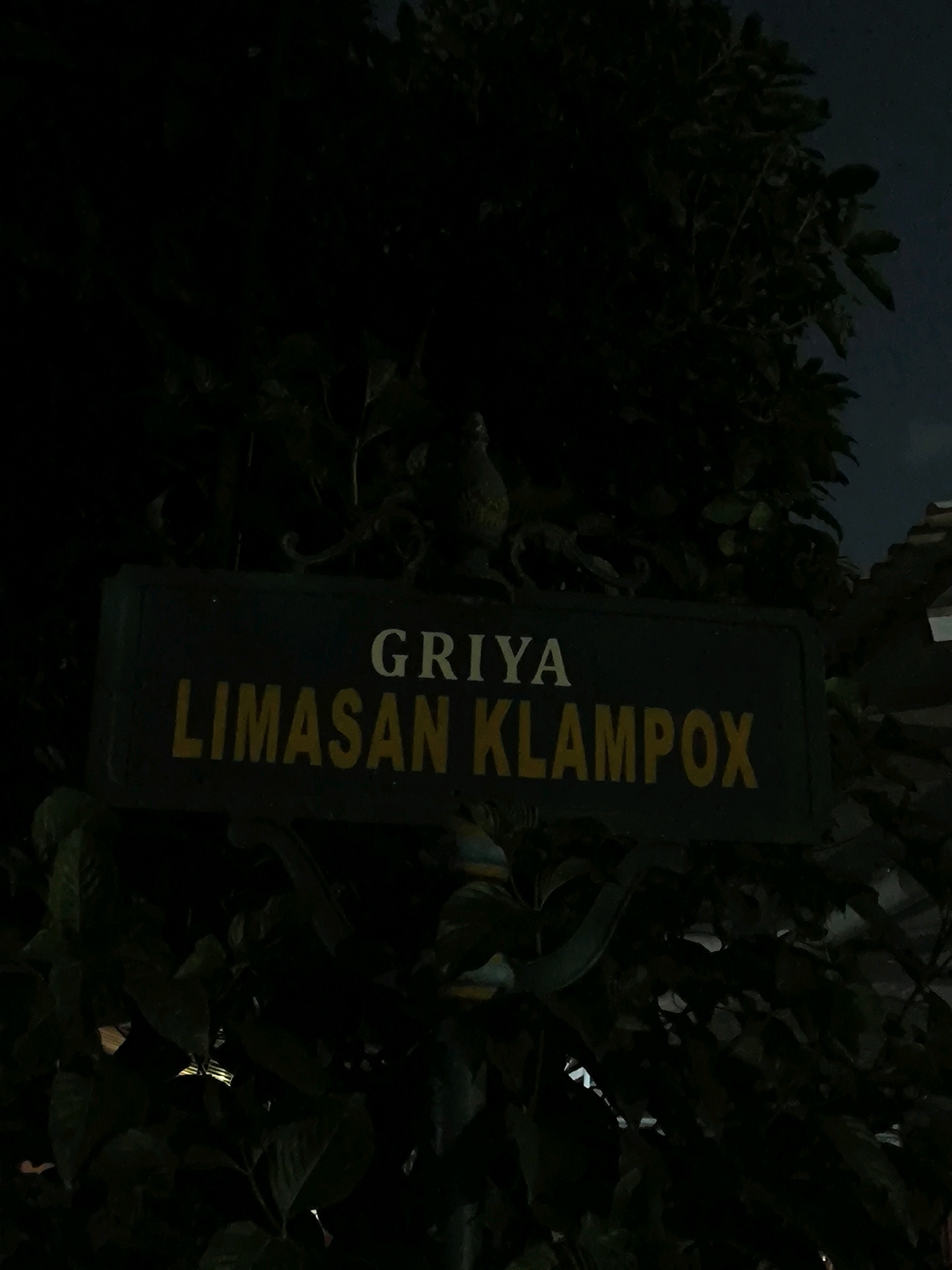 The image shows a dark, nighttime scene with a sign that reads "GRIYA LIMASAN KLAMPOX". The sign is dark green with white and yellow lettering. It is mounted on a dark pole and partially obscured by dark green foliage. The background is dark with some hints of the night sky and what appears to be a building. The scene is dimly lit, suggesting it is either nighttime or the area is not well-illuminated. There are no people or animals visible in the image.