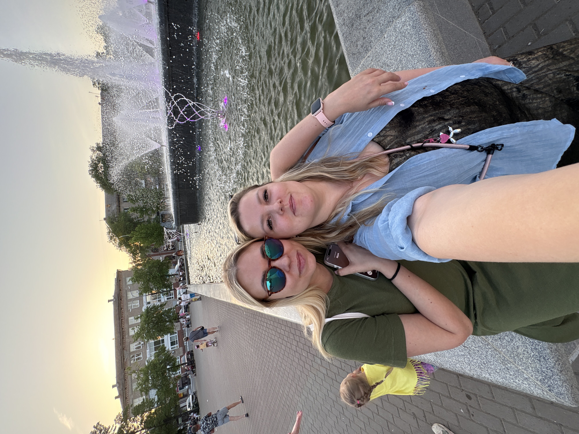 Two fair-skinned women pose closely for a selfie near a fountain in Chaikovsky, Russia. The woman in the foreground wears sunglasses and a green shirt, holding a phone in one hand. The other woman wears a blue shirt and a pink smartwatch. A fountain is visible in the background, with water jets and colorful lighting. In the distance, a brick walkway is populated with people and lined with trees and buildings. The sky suggests either sunrise or sunset.