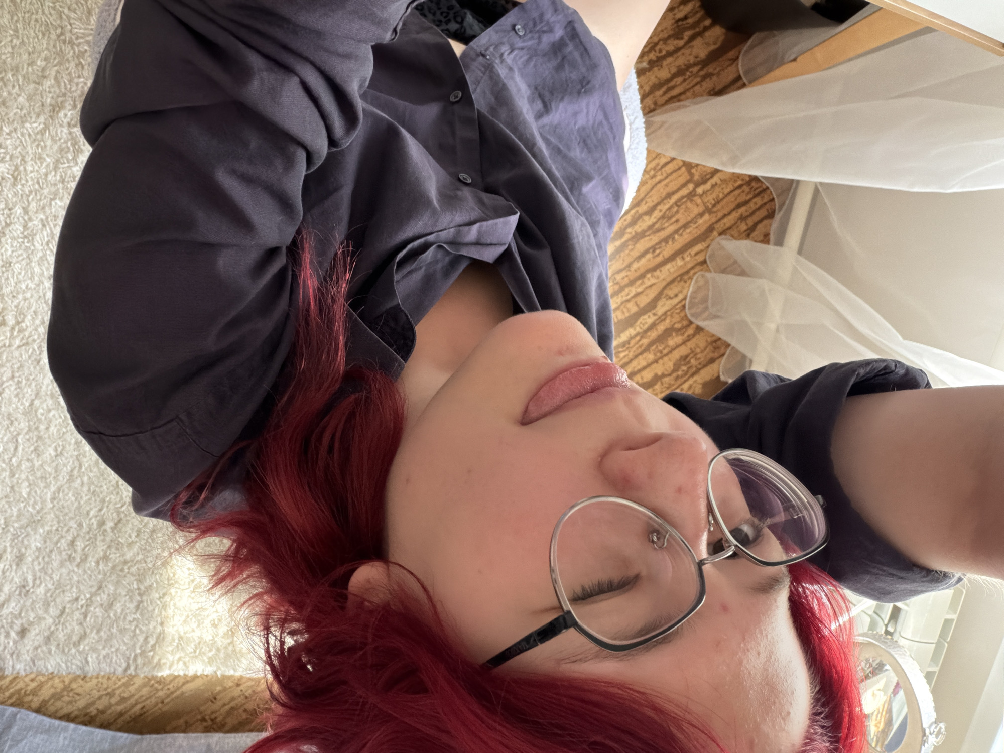The image shows a young person with bright red hair wearing glasses and a dark shirt. They are looking towards the camera with their tongue sticking out slightly and one eye closed. The background includes a white carpet, a wooden floor, and what appears to be white fabric. The lighting suggests an indoor setting, possibly a bedroom or living area. There are no discernible landmarks or clues that would indicate a specific location within Chelyabinsk, Russia.