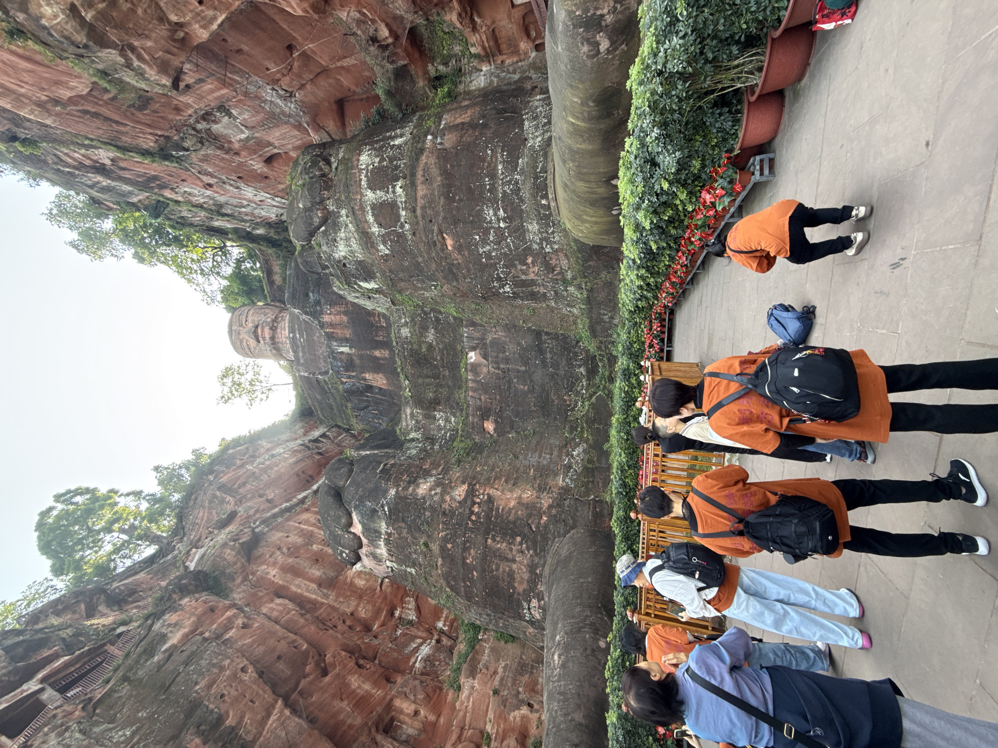 The image shows the Leshan Giant Buddha, located in Shizhong District, Leshan, Sichuan, China. The Buddha is carved into a cliff face. A line of tourists stand on a walkway next to a railing with potted plants, looking upwards at the statue. One person kneels near the edge of the walkway.