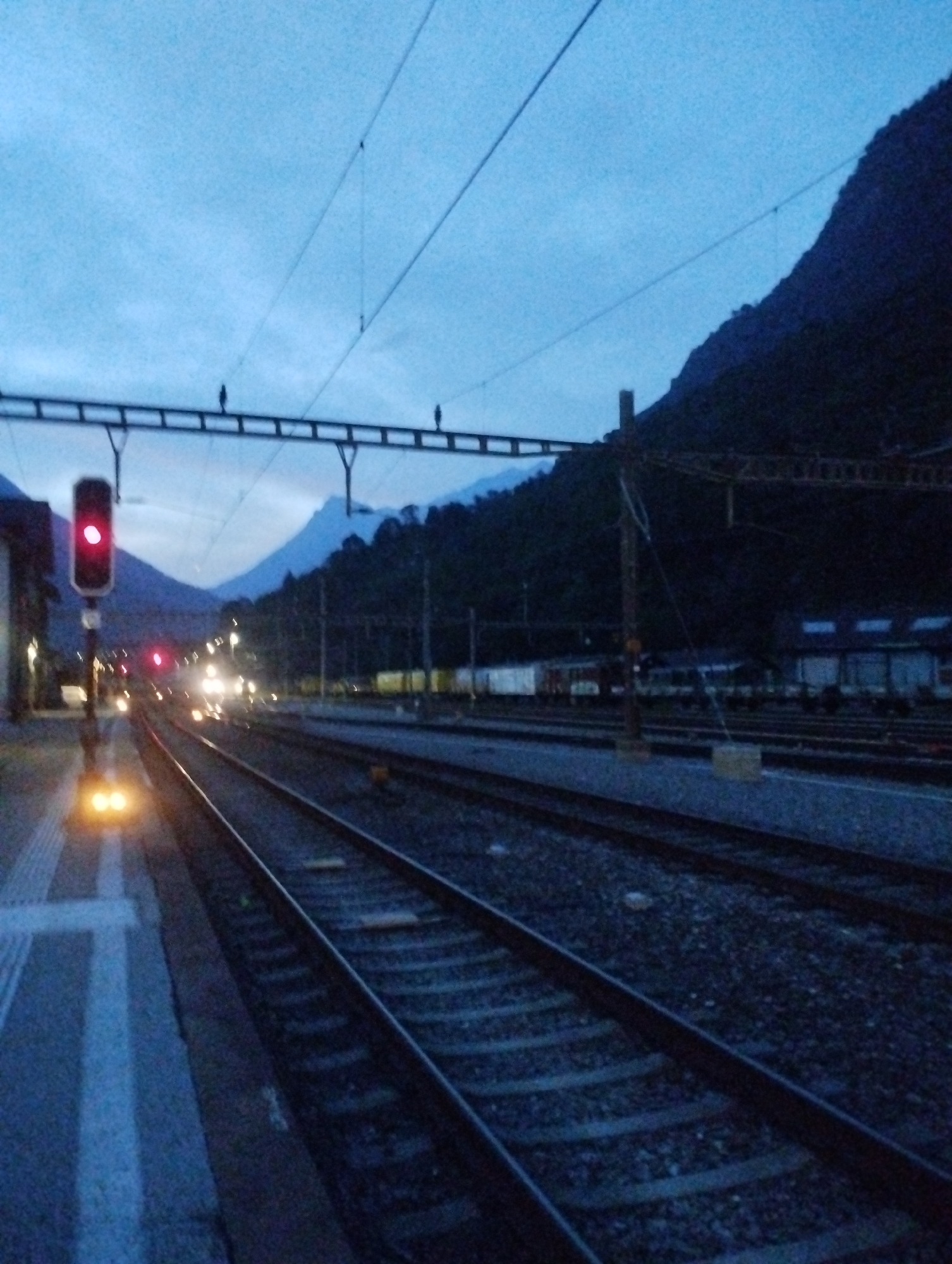The image captures a twilight scene at a train station in an unspecified location. In the foreground, train tracks stretch into the distance, bordered by a platform on the left. A red train signal is illuminated, indicating a stop. Further down the tracks, the headlights of an approaching or stationary train are visible. The background features a dark, mountainous landscape partially obscured by trees and overhead power lines for the trains. Buildings, possibly part of the station infrastructure, are visible to the right. The sky is a deep blue, with subtle cloud formations. The overall impression is one of a quiet, nocturnal or early morning atmosphere at a railway hub.