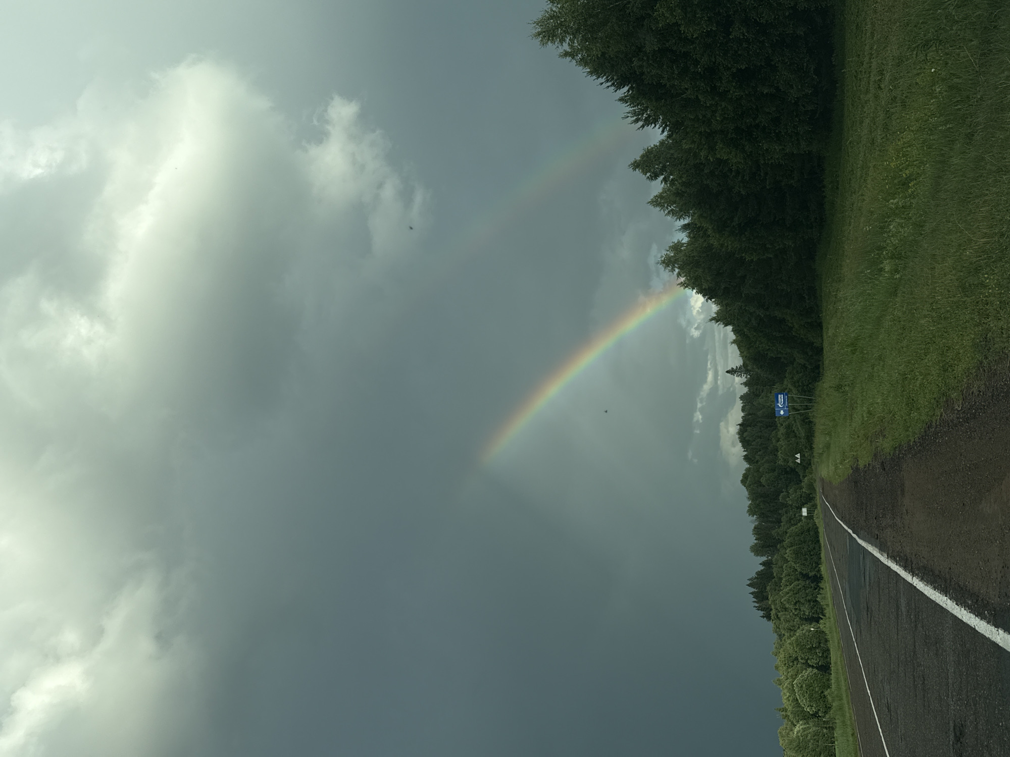 A double rainbow is visible in a dark, cloudy sky over a rural landscape in Кватчи, Russia. A paved road with a white line marking the shoulder runs from the lower left towards the distance. Lush green trees line the right side of the road, extending into a dense forest. The ground on the right is covered in green grass and some wildflowers. A blue road sign with a Gazprom logo and an arrow pointing to the left is visible near the edge of the trees. The sky is heavily clouded with a mix of dark storm clouds and lighter, brighter areas where the sun is breaking through, illuminating the rainbows.