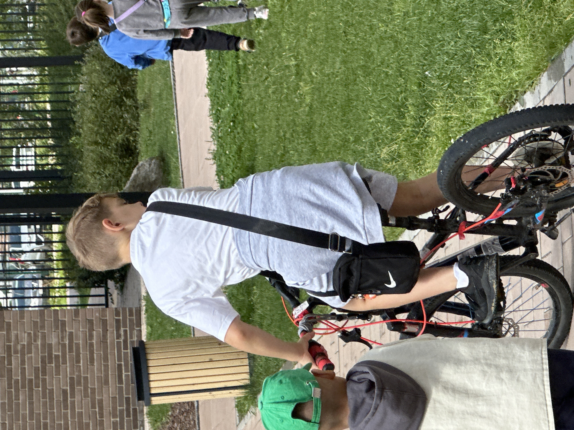 The image depicts an outdoor scene in a residential area, possibly Znamya Oktyabrya, Russia. A young boy with short blonde hair, wearing a white t-shirt and grey shorts, is positioned on a bicycle, seen from the rear and slightly to the side. He has a black shoulder bag with a white Nike logo slung across his chest. Another child, wearing a green cap and a light-colored hooded jacket, is partially visible in the foreground to the left, facing away from the viewer. In the background, two other children are walking on a paved path; one wears a blue jacket and the other grey sweatpants and a grey top. The environment includes a brick building on the left, a wooden trash receptacle, a black metal fence, lush green grass, and a paved walkway. The lighting suggests it is daytime.