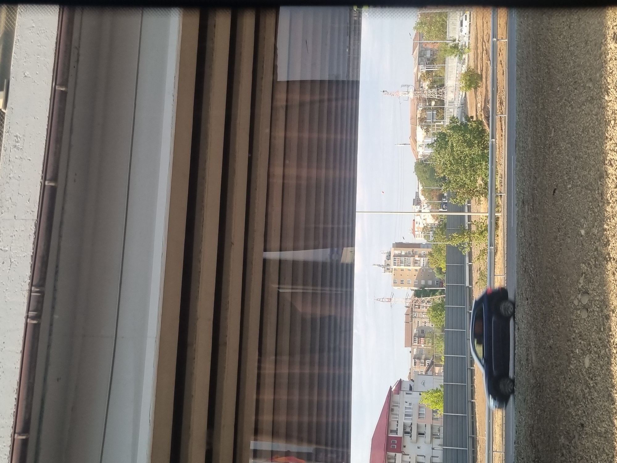 The image depicts a view through a partially open window or barrier, looking out onto a road and buildings. In the foreground, a dark-colored SUV is in motion on a highway, its wheels blurred due to speed. The road is bordered by a gravel shoulder and a metal guardrail. Beyond the road, a sound barrier is visible, with buildings of various heights and architectural styles situated in the background. These buildings appear to be a mix of residential and commercial structures, with some featuring red roofs. Trees and vegetation are interspersed among the buildings. A prominent communication tower is also visible in the middle ground. The sky is overcast, suggesting a cloudy day. The scene appears to be an urban or suburban environment, likely in Oradea, Romania, given the context. The perspective suggests the photograph was taken from an elevated position, possibly from a bridge or a higher floor of a building.