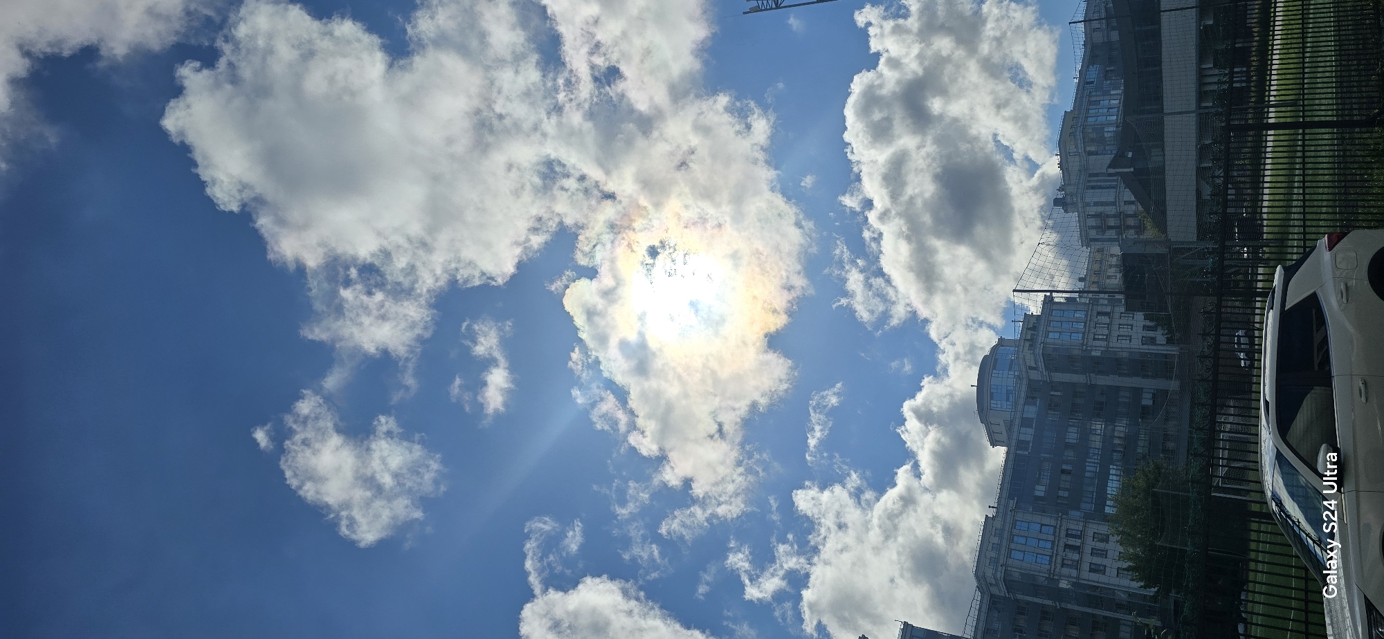 The image is a vertical, full-frame shot of a bright, partly cloudy sky over an urban area, likely Saint Petersburg, Russia. The sun is partially obscured by clouds, creating a colorful iridescence and sunbeams. In the foreground, the upper part of a white SUV is visible, with the text "Galaxy S24 Ultra" at the bottom left. Behind the car is a black metal fence, beyond which is a green field, possibly a sports ground, with a soccer goal visible. Further in the background, a row of modern apartment buildings with numerous windows stretches across the frame. The sky is a vibrant blue with large, fluffy white clouds.