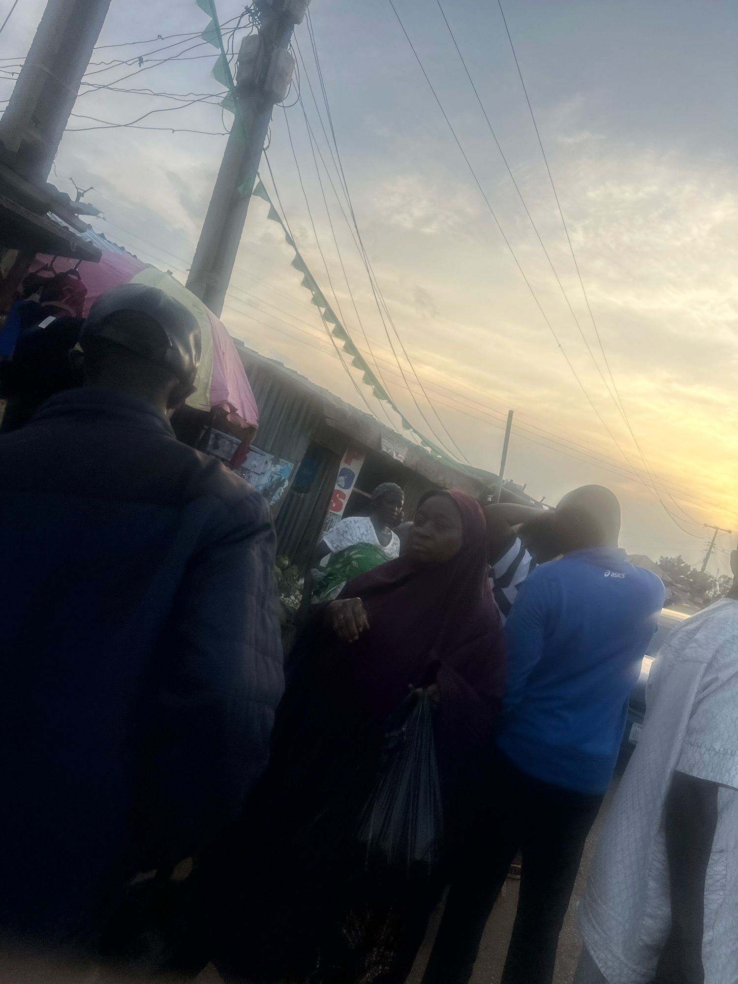 The image captures a street scene in Gura Top, Nigeria, characterized by a mix of informal market stalls and urban infrastructure. A prominent utility pole with numerous wires and green triangular flags strung along it dominates the center-left of the frame. To the left of the pole, corrugated metal structures with hanging fabrics suggest market stalls.
In the foreground and mid-ground, several individuals are visible. On the far left, a person wearing a dark jacket and a cap is seen from behind, facing away from the camera. To their right, a woman in a maroon hijab and a dark headscarf holds a black plastic bag. Further to the right, a man in a blue long-sleeved shirt with "Asics" branding is visible from the back. Partially obscured behind him, another person in white clothing is seen from the right side. In the background, more individuals and market structures are present, including what appears to be a sign with the letters "POS."
The sky above is filled with soft clouds and a warm glow, indicating either sunrise or sunset, with power lines crisscrossing the expanse. The overall impression is one of a bustling, everyday urban environment, likely a market area, at a transitional time of day.