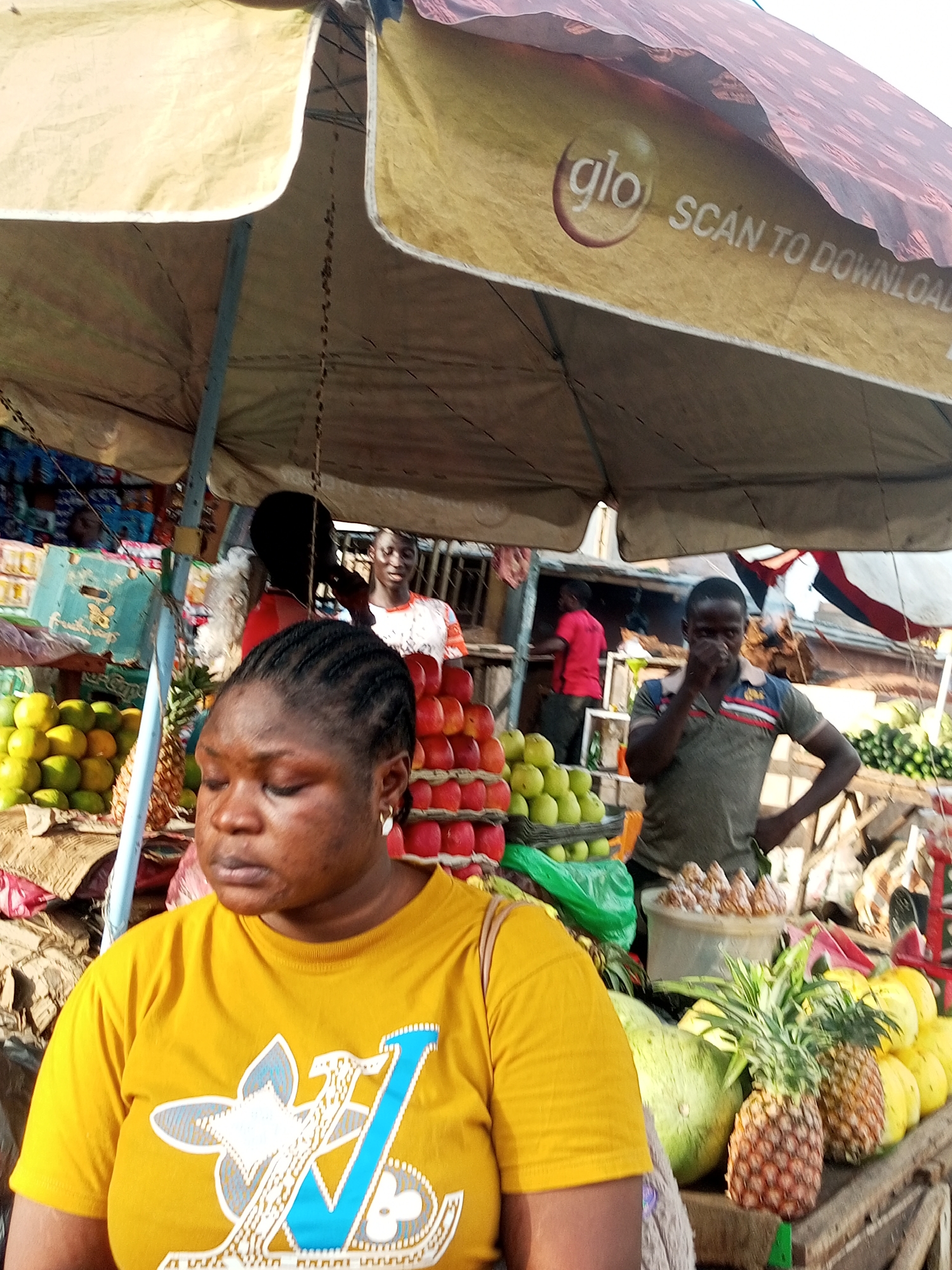 This is a medium shot of a street market scene, likely in a tropical or subtropical region, given the abundance of fruit. In the foreground, a woman with braided hair wearing a yellow t-shirt with a large blue and white graphic is facing slightly away from the camera, her expression somewhat somber. Behind her, a stall displays a variety of fruits, including stacks of red and green apples, pineapples, and what appear to be oranges or mangoes. Further in the background, several other individuals are visible, some interacting with the market stalls. One man in a grey and striped polo shirt is standing with his hand near his face. Another person in a red shirt is visible further back. The scene is covered by a large, weathered awning, likely for shade. The awning has a logo and text that reads "glo scan to download". The time of day appears to be daytime, and the weather seems bright and possibly warm, suggesting an outdoor market environment.