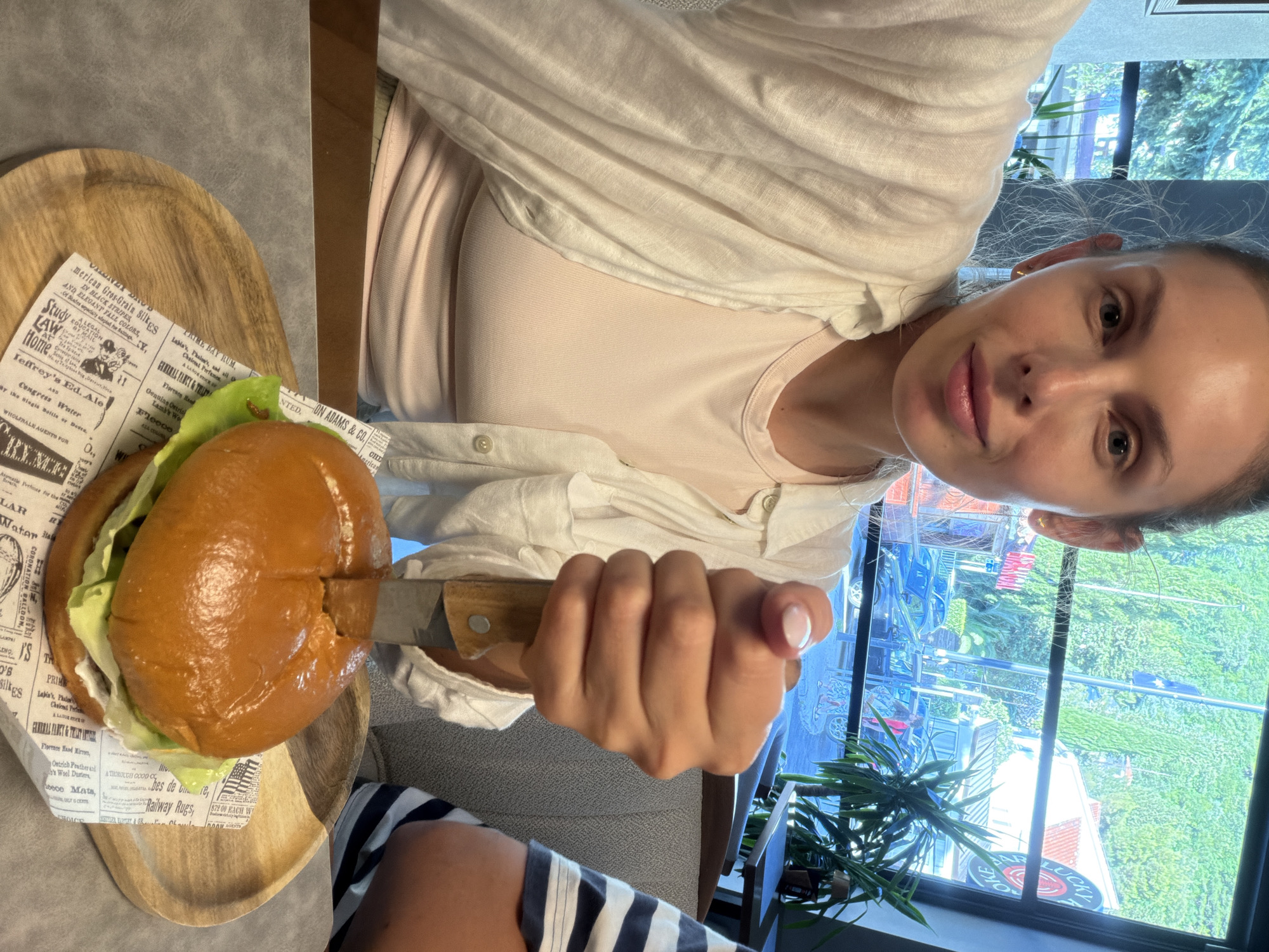 A woman with blonde hair pulled back in a bun is taking a selfie while holding a knife embedded in a hamburger. She is wearing a light pink tank top and an unbuttoned white linen shirt. The hamburger is on a wooden plate with a piece of newspaper underneath it, which is placed on a grey table. In the background, a large window shows a green hillside with trees and buildings. The setting appears to be a restaurant or cafe in Budva, Montenegro, suggested by the architecture and lush greenery visible outside.