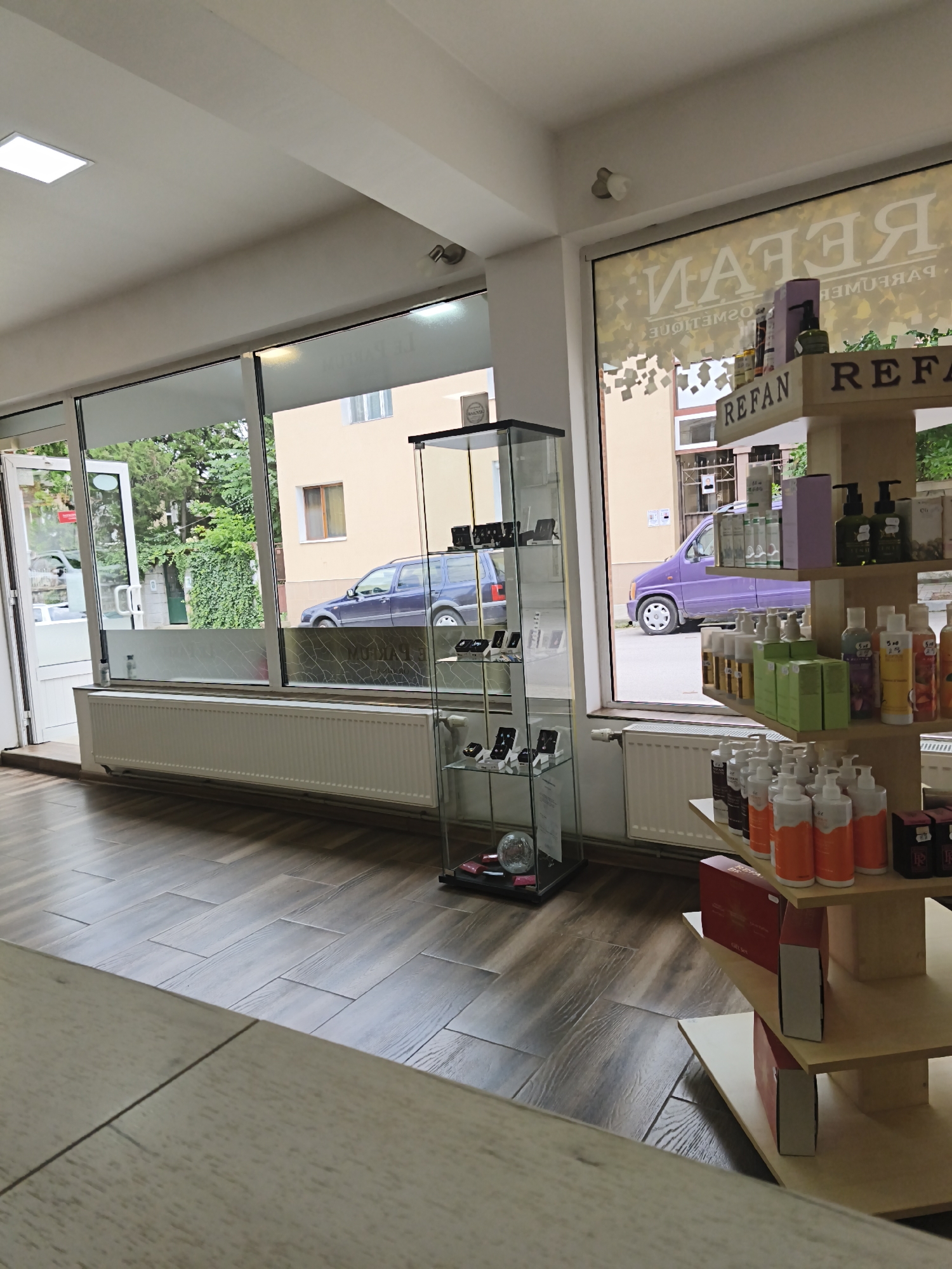 The image depicts the interior of a Refan perfume and cosmetics store located in Harmanli, Bulgaria. The store features large windows offering a view of the street, where a purple car is parked. Inside, the store is organized with display shelves showcasing various perfume bottles, lotions, and gift sets. A glass display cabinet in the foreground holds what appear to be jewelry or accessories. The floor is tiled with a wood-like pattern, and a white radiator runs along the bottom of the windows. The lighting appears to be a mix of natural light from the windows and interior store lighting. There are no people visible in the image.