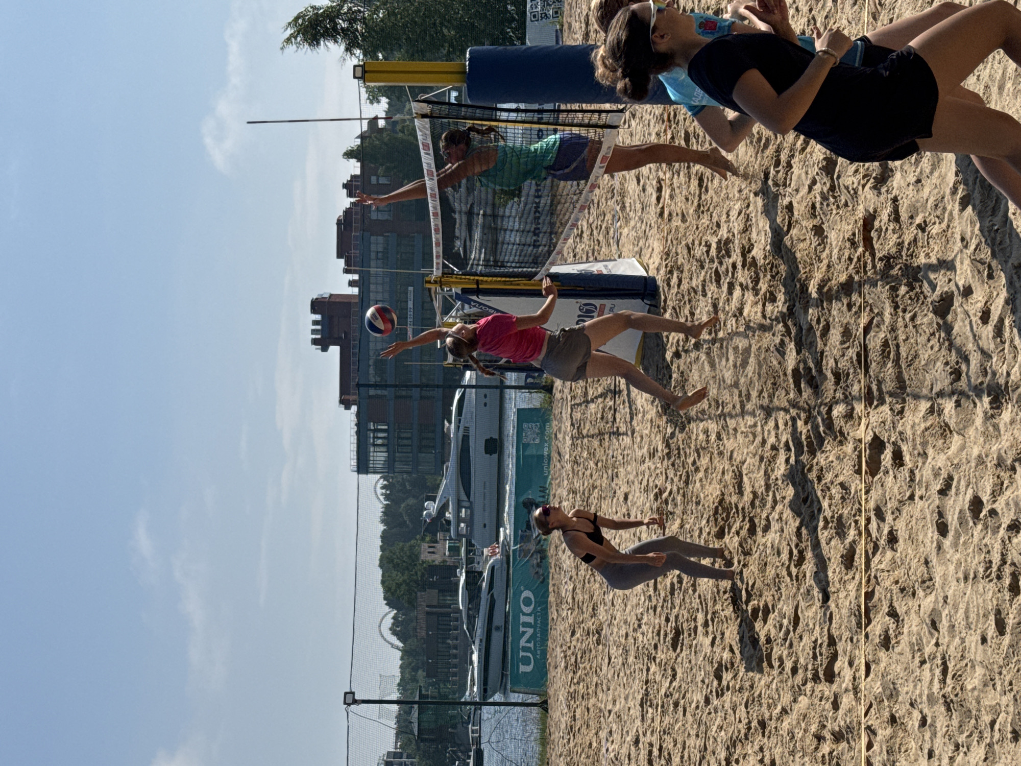 This media file shows a beach volleyball game with several people actively playing on a sandy court. The game appears to be taking place on a sunny day, as indicated by the bright lighting and shadows. In the background, there are large buildings, boats docked in a body of water, and trees, suggesting an urban or semi-urban waterfront location. One of the players is in mid-air, reaching to hit the volleyball. Another player is on the ground, diving to save the ball. A third player is watching the game. The net is taut and set up for the game. The scene evokes a sense of athletic competition and recreation. Visible text includes "UNIOWEX.COM" on a banner and "UNIO" on another banner. The image is oriented vertically.