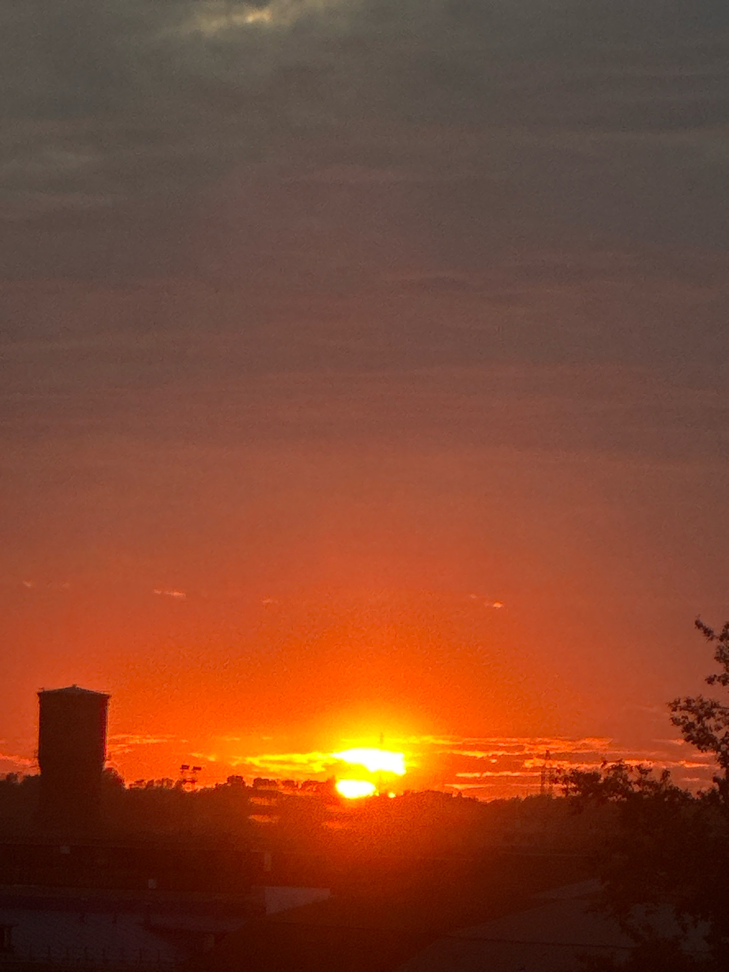 The image captures a sunset over Biysk, Russia. A dark, cylindrical water tower stands silhouetted against the vibrant orange and red sky on the left side of the frame. The sun, a bright orb with a halo of light, is setting behind a low, indistinct treeline and structures. Wispy clouds are scattered across the sky, tinged with the colors of the sunset. The foreground is dominated by dark silhouettes of buildings and trees. The overall mood is tranquil and dramatic, characteristic of a sunset.