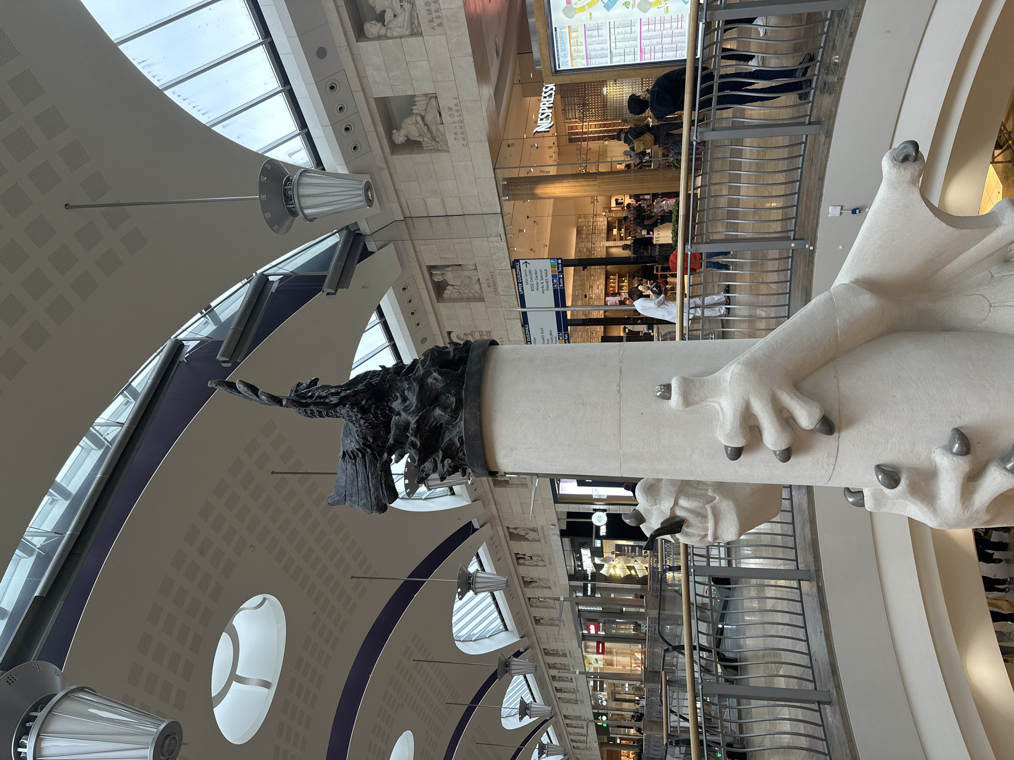 Here is a detailed description of the image:
The image depicts the interior of a shopping center in the City of London, United Kingdom, specifically the One New Change shopping center near St. Paul's Cathedral.
The scene is focused on a large sculptural column adorned with a bronze eagle at the top and massive white dragon claws emerging from the sides. The column extends upward into the multi-level atrium of the shopping center.
Architectural details include a curved ceiling with skylights and evenly spaced, drum-shaped light fixtures suspended from the ceiling. Relief sculptures are visible on the walls of the upper levels, along with store signs, including a Nespresso shop.
People can be seen on the walkways of the upper levels, going about their business. The photo is taken from a slightly low angle, emphasizing the height of the column and the expansive space.