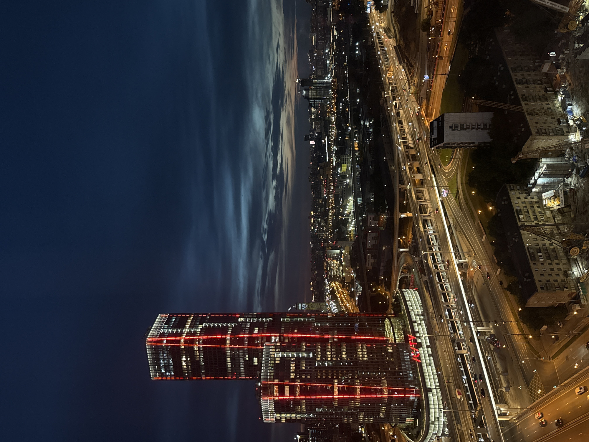 This is a night aerial view of the city of Khimki, Russia, showcasing modern skyscrapers and infrastructure. Two prominent skyscrapers, illuminated with vertical red lights, dominate the left side of the frame. The taller skyscraper is rectangular in shape, while the adjacent shorter one has a curved base. The word "IG/CITY" is clearly visible in red lettering on the lower building's facade. Surrounding these buildings is an extensive network of multi-level highway overpasses and roads, teeming with illuminated vehicles. In the distance, a cityscape of various buildings stretches towards the horizon, dotted with streetlights and building lights, under a dark, cloudy sky with a hint of twilight. The lower portion of the image reveals more urban elements, including apartment buildings, construction sites with cranes, and more roadways. The scene captures a vibrant, illuminated urban environment at dusk.