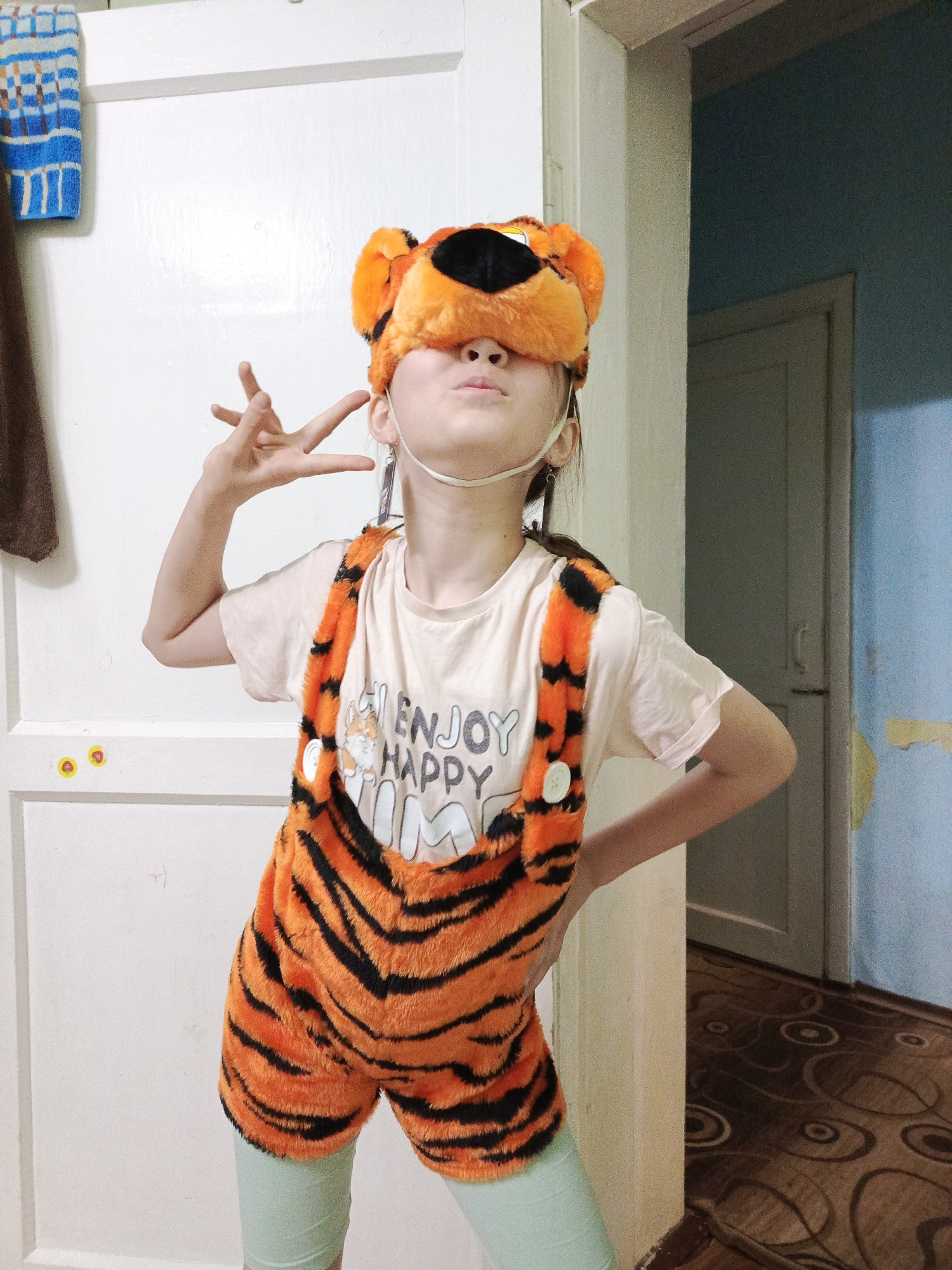 A young person, presumably a child, poses indoors wearing an orange and black tiger costume, consisting of a furry headpiece covering their eyes and tiger-striped overalls over a light-colored t-shirt. The individual makes a hand gesture while placing their other hand on their hip. The background includes a white door with a few yellow stickers, a doorway leading to a room with blue walls and another door, and part of a patterned rug. The environment is consistent with a domestic interior. This photo was taken in Garevaya, Russia.