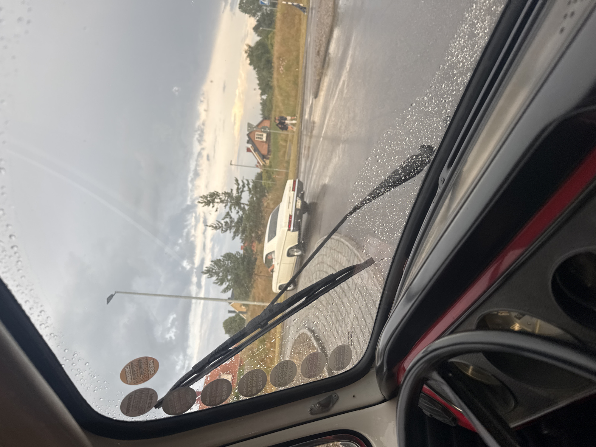 The image is taken from inside a vehicle looking out through a rain-streaked windshield. A white vintage car is driving away from the camera on a wet road. There are two people walking in the distance on the sidewalk. The environment appears to be a residential area with houses and trees visible. The sky is cloudy, suggesting inclement weather. There are several circular stickers on the lower left portion of the windshield, which appear to be vehicle inspection stickers. The car interior, including a steering wheel and dashboard, is visible in the foreground. The location is identified as Falkenberg, Sweden.