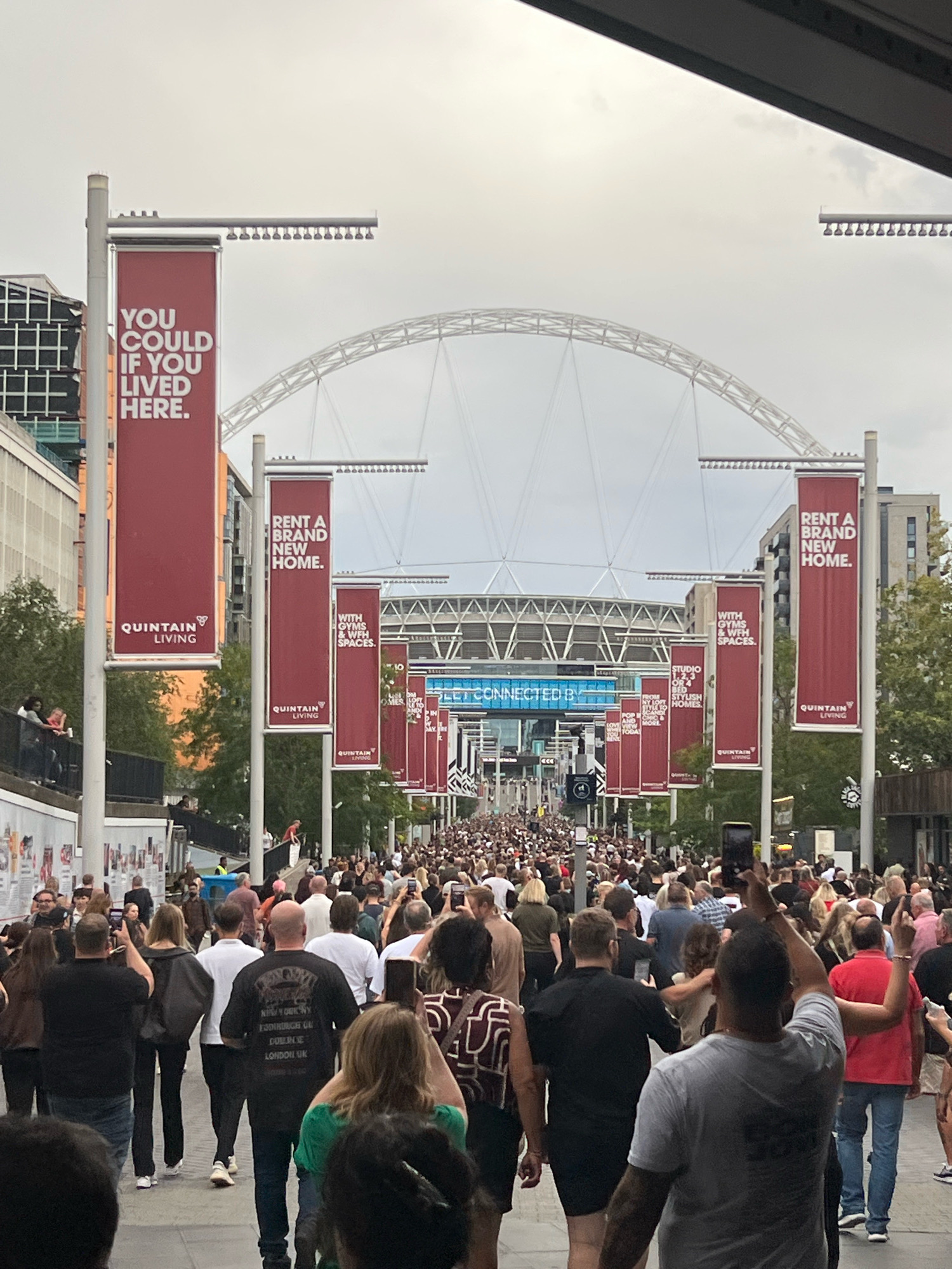 This is an outdoor, eye-level, medium-shot photograph taken during daylight under an overcast sky. The scene depicts a large crowd of people walking down a wide, paved walkway that leads towards the iconic Wembley Stadium in London, United Kingdom.
The walkway is flanked by lampposts on either side, each adorned with several large, maroon banners. These banners feature white text promoting "QUINTAIN LIVING" and advertising "RENT A BRAND NEW HOME," with slogans like "YOU COULD IF YOU LIVED HERE," "WITH GYMS & WFH SPACES," and "STUDIO, 1, 2 & 3 BED STYLISH HOMES." In the background, the distinctive arch of Wembley Stadium is visible. The crowd is densely packed, with individuals of diverse ages and ethnicities walking in the same direction. Many people are holding up mobile phones, presumably to take pictures or videos of the event or the stadium. The overall atmosphere appears to be one of a public gathering, possibly before or after an event at the stadium.