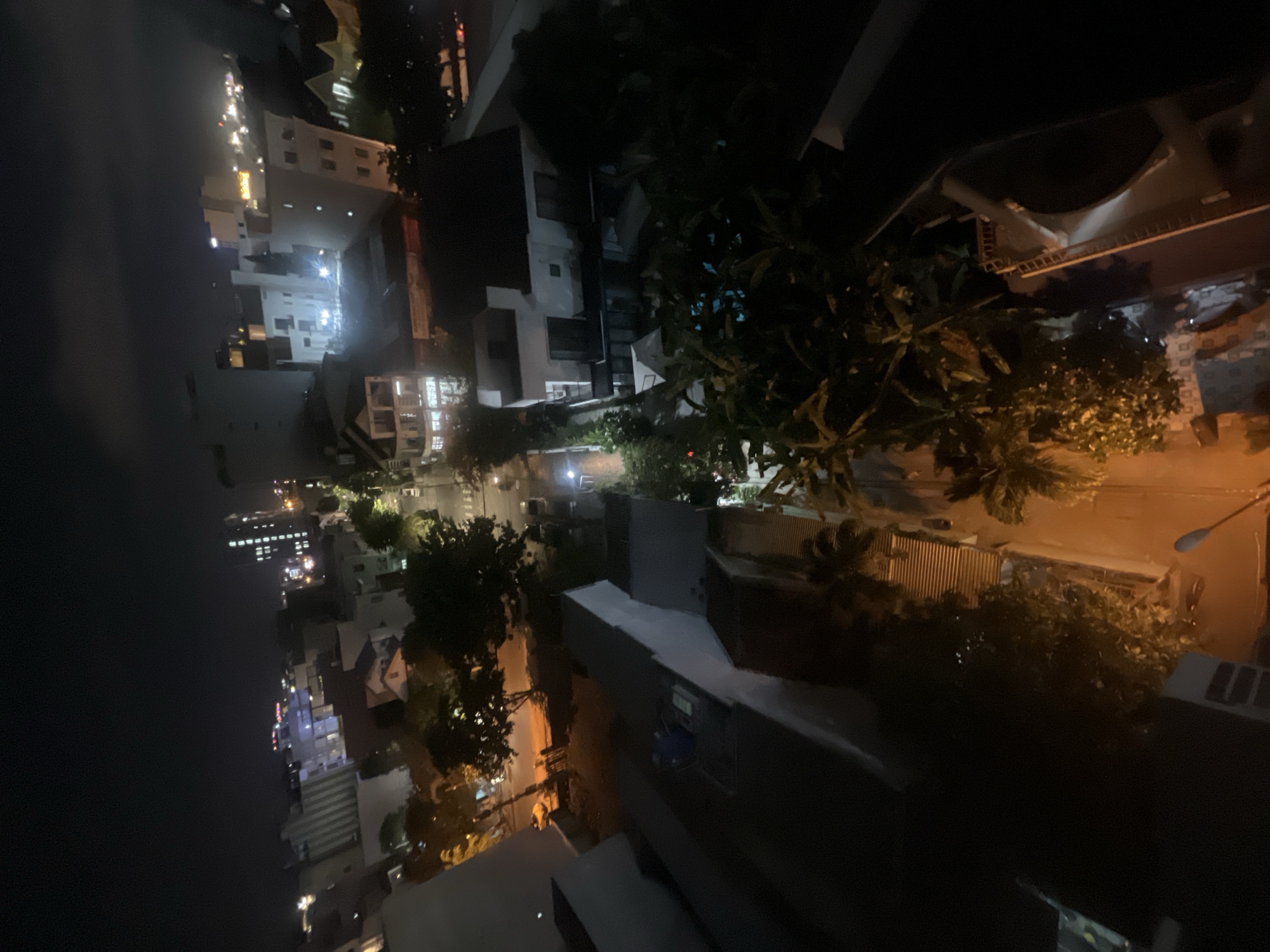 The image is a nocturnal, high-angle view of a city street scene in Da Nang, Vietnam. Buildings of varying heights, many with illuminated windows, line the street. Trees with dark foliage are interspersed among the structures, some appearing to be quite large and leafy. The street itself is visible in patches, with car headlights and streetlights casting a warm, orange glow on the wet pavement, suggesting recent rain or high humidity. The overall impression is of a vibrant urban environment at night, with the city lights reflecting on the surfaces. The perspective suggests the photo was taken from a higher vantage point, possibly a balcony or another building. There is no discernible text in the image.