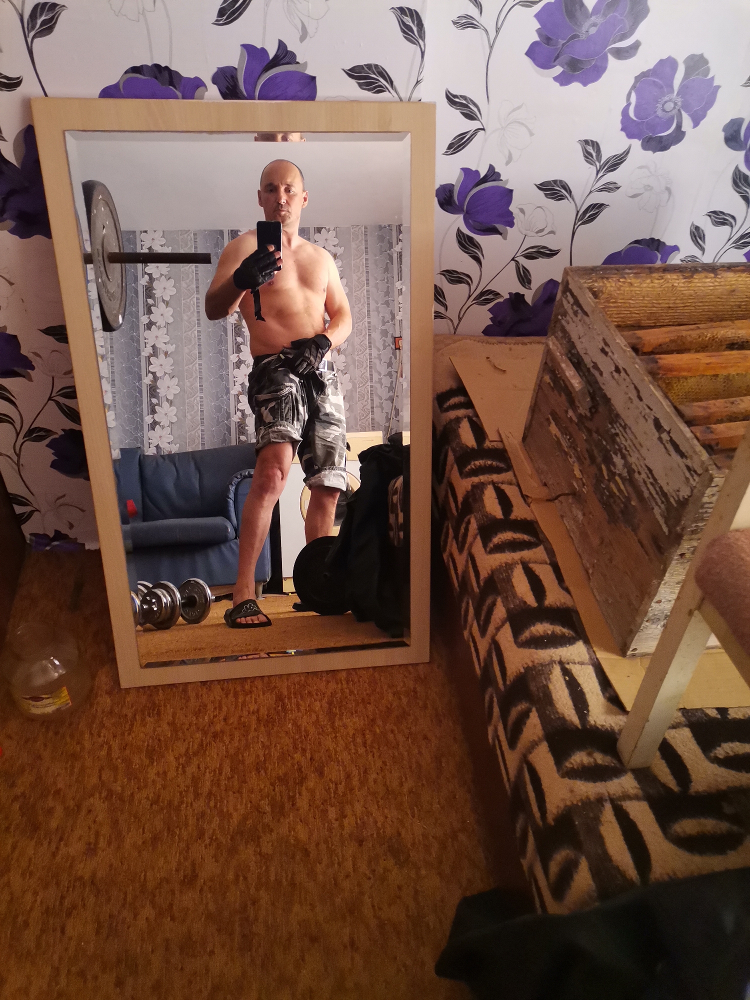 A shirtless man is shown reflected in a mirror. He is holding a phone in his left hand and wearing shorts, black gloves, and sandals. The background visible in the mirror reveals a barbell with weights, dumbbells on the floor, and a blue sofa against a patterned wallpaper. The environment outside the mirror includes a patterned couch or bedding and a wooden object that appears to be furniture or a frame. The location is Unknown, Unknown.