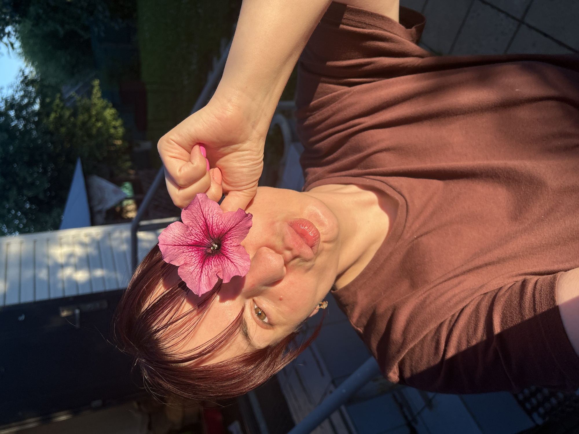 A person is outdoors in what appears to be a backyard or patio area, holding a pink petunia flower to their face. The person, with reddish-brown hair, is looking towards the camera with their eyes slightly open and lips puckered. The flower is placed near their eye. They are wearing a brown t-shirt. The lighting suggests it is daytime, possibly late afternoon, due to the warm, angled sunlight creating shadows. In the background, there are trees, a fence or railing, and some roofing, indicating a residential setting. The sky is visible, appearing clear and blue. The overall mood seems playful or whimsical. No visible text is present in the image.