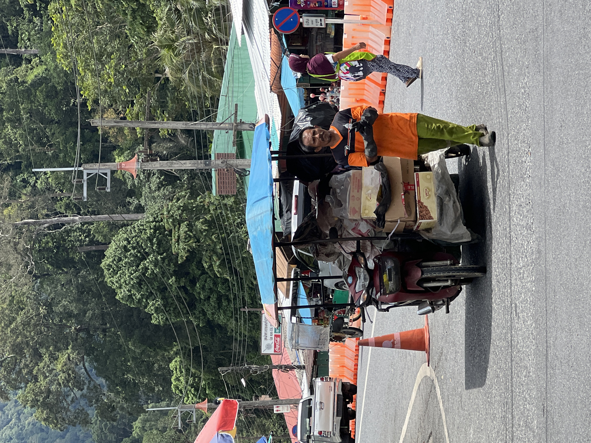 This media file depicts a street scene in Tambon Wang Prachan, Thailand. The primary subjects are two individuals, likely a vendor and a customer, interacting near a cart laden with goods. The vendor, a man wearing an orange shirt and green pants, stands next to the cart. He is wearing dark gloves and appears to be holding something. The cart itself is covered with a blue tarp and is filled with boxes and bags, suggesting it is a mobile stall or a delivery vehicle. In the background, another person, possibly a customer, is visible wearing a purple headscarf and a colorful shirt, standing behind orange traffic barriers.
The scene appears to be set in a bustling outdoor market or roadside. The presence of overhead power lines, lush green trees in the background, and the colorful awnings of nearby stalls indicate a tropical environment. The weather seems fair, with bright sunlight casting shadows on the asphalt road, suggesting it is daytime. The overall atmosphere seems to be that of commerce and daily life, with people engaged in transactions or preparing for them.
Visible text includes a Coca-Cola logo on a sign attached to one of the stalls. Other signs are present but too blurry to read. The road has painted white lines, and orange traffic cones are placed along the side, possibly indicating a temporary closure or a designated area. The presence of a red motorcycle attached to the cart suggests it is the mode of transport for the vendor's goods.