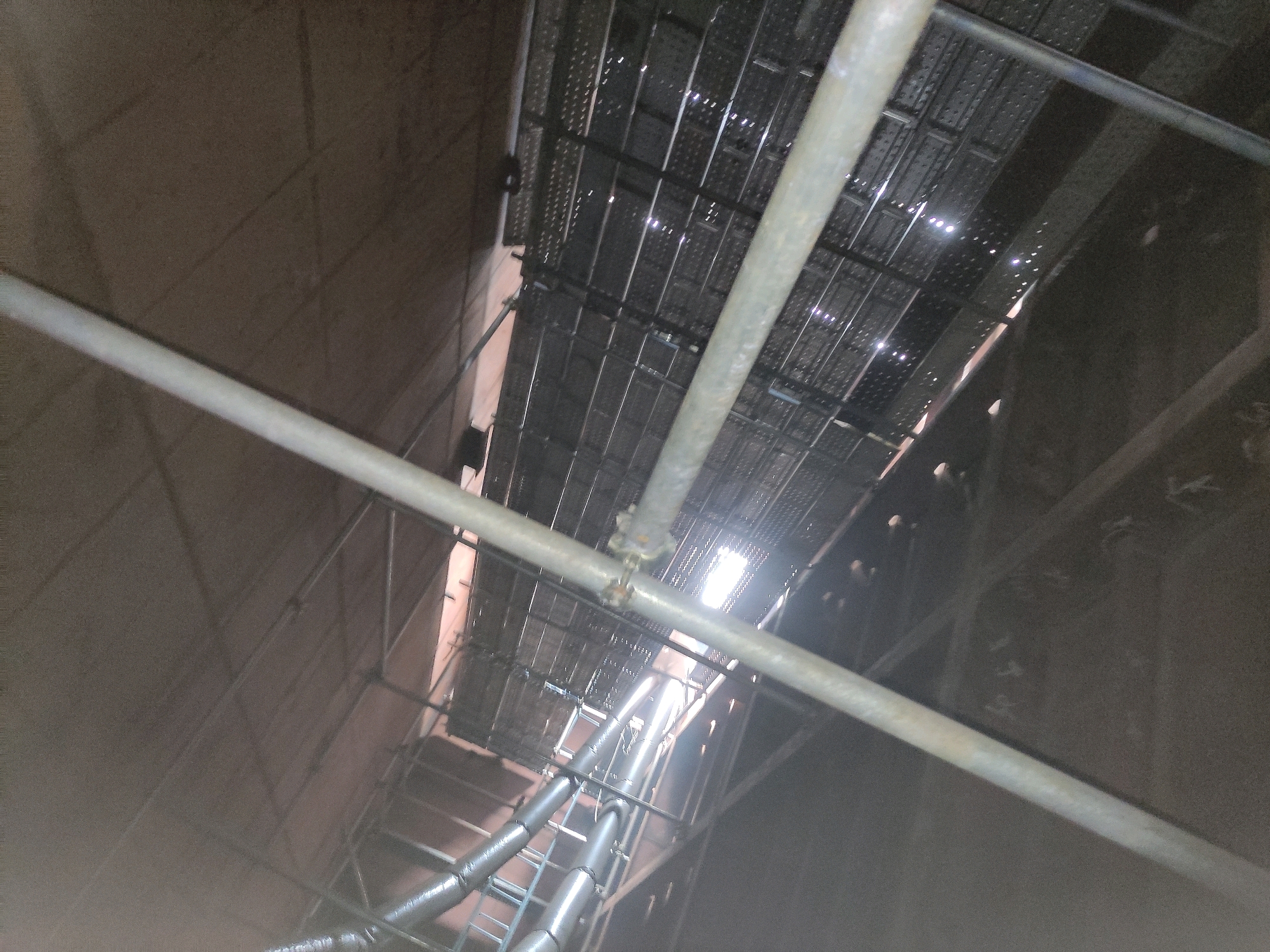 This image shows an interior view of a large, unfinished industrial structure, likely a tank or silo, in Batam, Indonesia. The perspective is looking upwards, revealing multiple levels of scaffolding and metallic grating. Several large diameter pipes, some with insulation, are visible running horizontally along the lower sections of the structure. The walls of the structure are a rough, textured concrete or similar material. Natural light is entering from an opening at the top of the structure, illuminating dust particles in the air. The overall scene suggests ongoing construction or maintenance work.