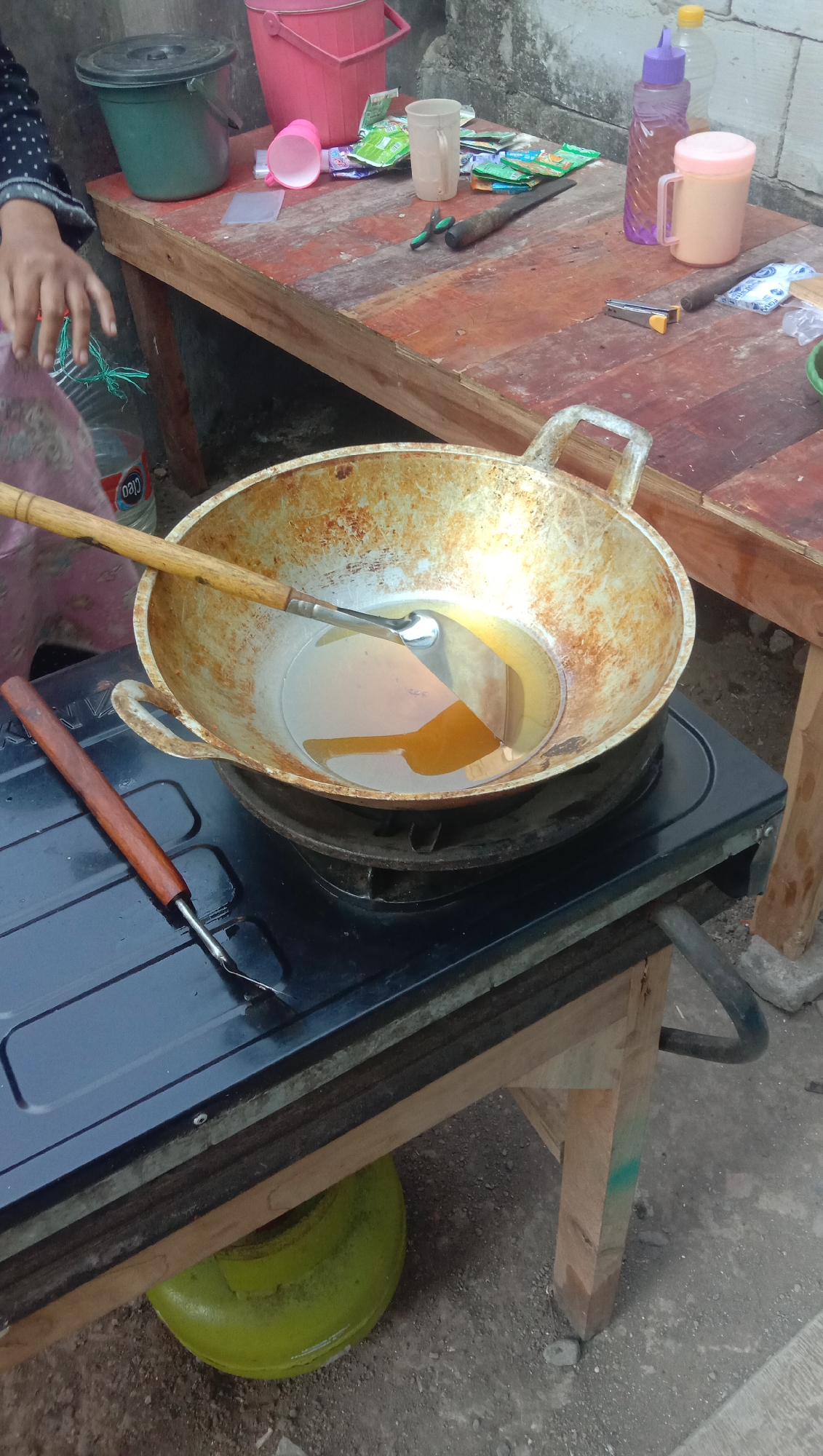 The image depicts an outdoor cooking scene, likely in Karang Anyar, Indonesia. A gas stove with a wok filled with hot oil is the central focus. The wok is a well-used, metallic pan with a light-colored handle. A metal spatula with a wooden handle rests in the wok. Beside the stove, a wooden table is laden with various items, including a pink bucket, a green bucket with a lid, several packets of what appear to be food seasonings or instant noodles, a clear plastic cup, a purple water bottle, and a light orange mug with a lid. Also on the table are a pair of small pliers, a knife, and a silver stapler. A person's hand and arm are visible on the left, partially obscured, suggesting they are preparing or overseeing the cooking. The background shows a textured wall and the ground is dirt and gravel. A green gas cylinder is visible beneath the stove. The lighting suggests it is daytime.