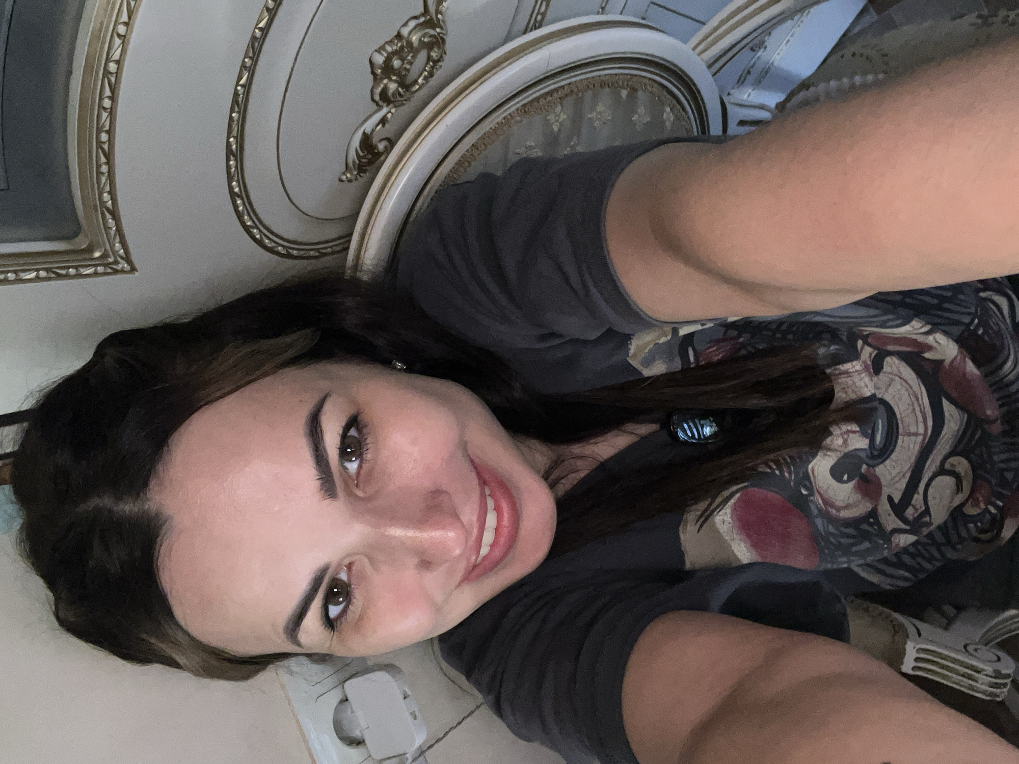 A woman with long, dark brown hair smiles and looks directly at the camera in a close-up selfie. She is wearing a dark t-shirt with a graphic print. Her left arm is extended, holding the camera. Behind her, to her right, is an ornate, white and gold chair. To her left, a wall with a light-colored outlet is visible. The lighting appears to be natural, coming from the left. The scene is indoors.