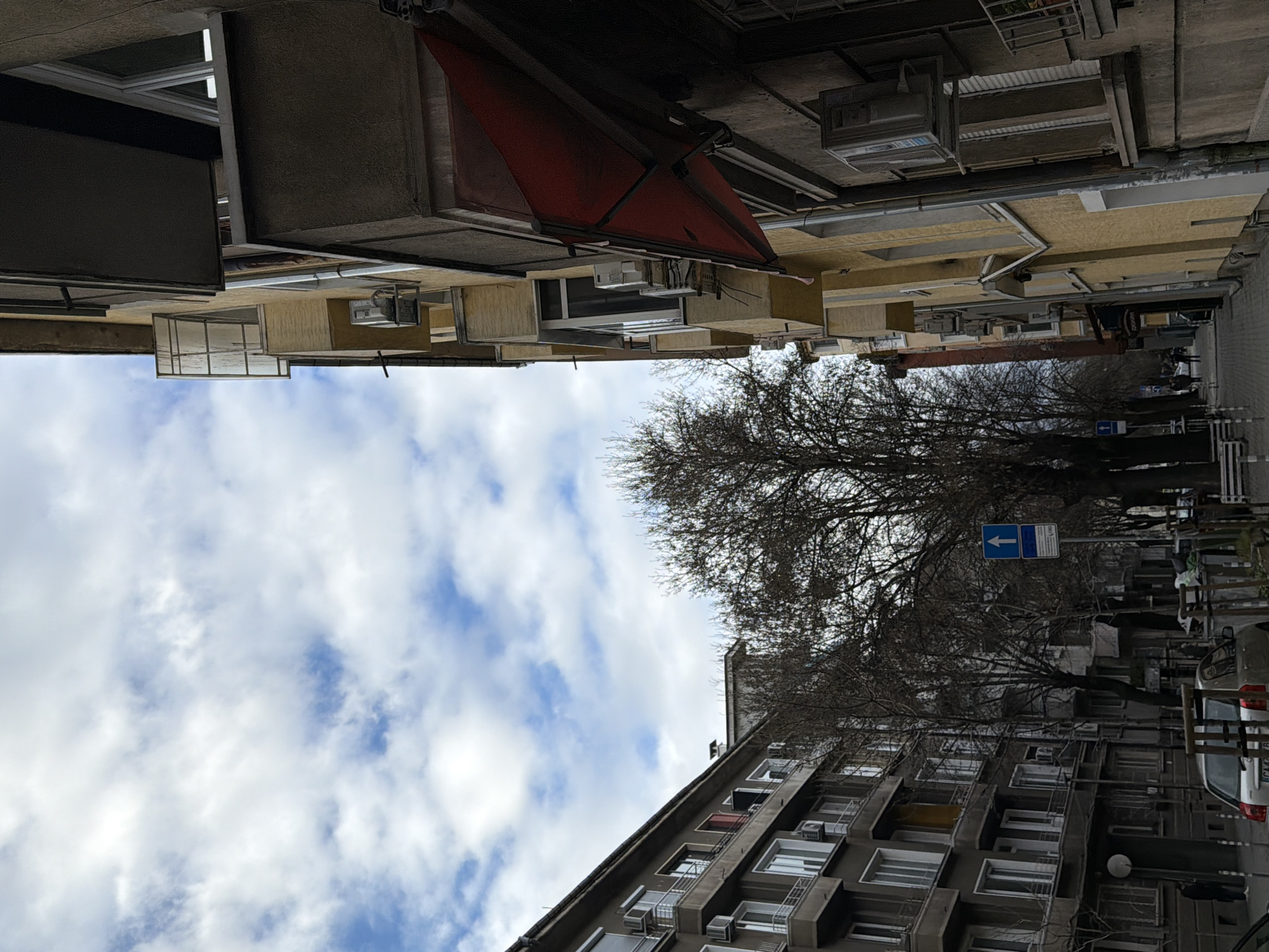 The image depicts a street scene in Sofia, Bulgaria, viewed from a low angle looking upwards. On either side of the street are buildings, mostly residential apartment blocks. The building on the right has numerous windows and balconies. On the left side of the street are buildings with yellow paint. A red awning is visible on the upper-left side of the image. Bare trees line the street, partially obscuring the buildings. A street sign with a blue arrow pointing left is present on the right side of the street. Cars are parked along the street. The sky above is cloudy with patches of blue.