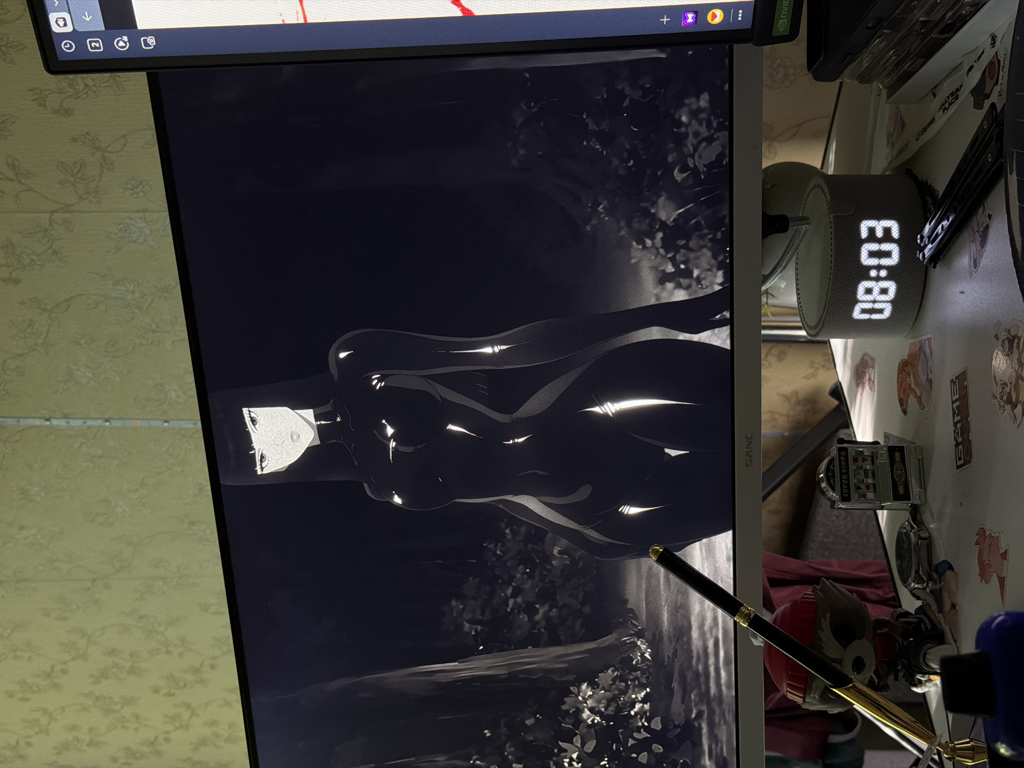 The image displays a computer monitor and a desk cluttered with various objects. On the monitor, an anime-style illustration is visible, featuring a black and white depiction of a female figure with striking highlights, suggesting a high-contrast aesthetic. The background of the illustration appears to be a shadowy forest or garden setting. The monitor itself is positioned on a desk.
To the right of the monitor, on the desk, an alarm clock displays the time "08:03" in a bright, digital font. Also on the desk, there are several small objects including what appears to be a miniature slot machine, a watch, and figurines. A black pen with gold accents is also visible, resting in a way that suggests it might be used for writing or drawing. The overall impression is that of a personal workspace. The wallpaper in the background of the room has a floral pattern. The image is likely taken indoors, in a room that is somewhat messy but organized for personal use. The time of day indicated by the clock is early morning.