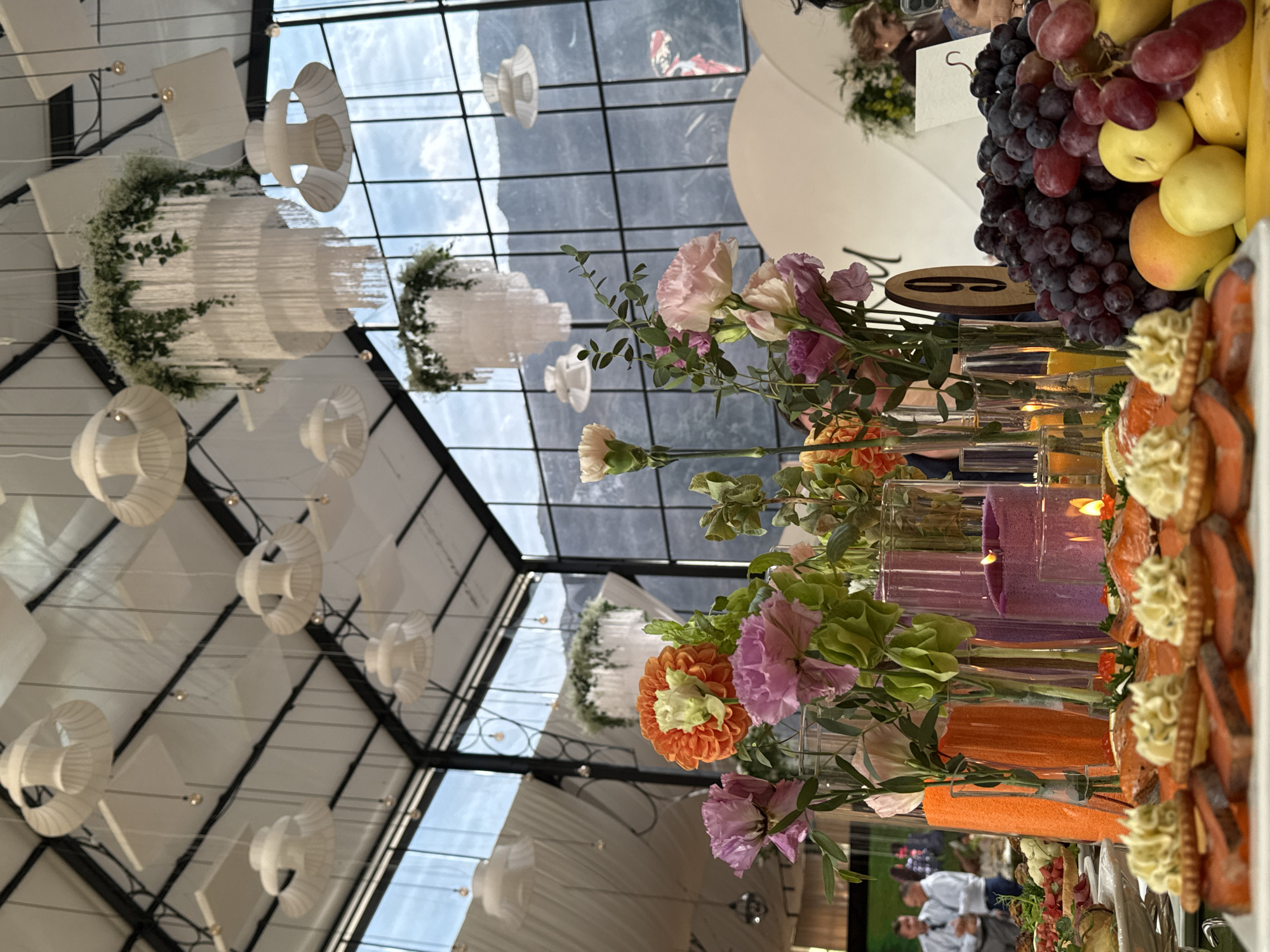 The image depicts a catered event at a venue with a high ceiling and glass walls, offering a view of mountains. The scene focuses on a table laden with food and floral arrangements. In the foreground, there are various appetizers, including small bites topped with creamy spread and slices of salmon. Behind these, a vibrant display of flowers in glass vases adds color and elegance. Several candles are lit, casting a warm glow on the table. In the background, guests are visible, some mingling and others seated. Large, decorative chandeliers hang from the ceiling, complementing the overall festive atmosphere. The venue appears to be a modern banquet hall or event space, possibly located in or near Arashan, Kyrgyzstan, given the mountain backdrop.