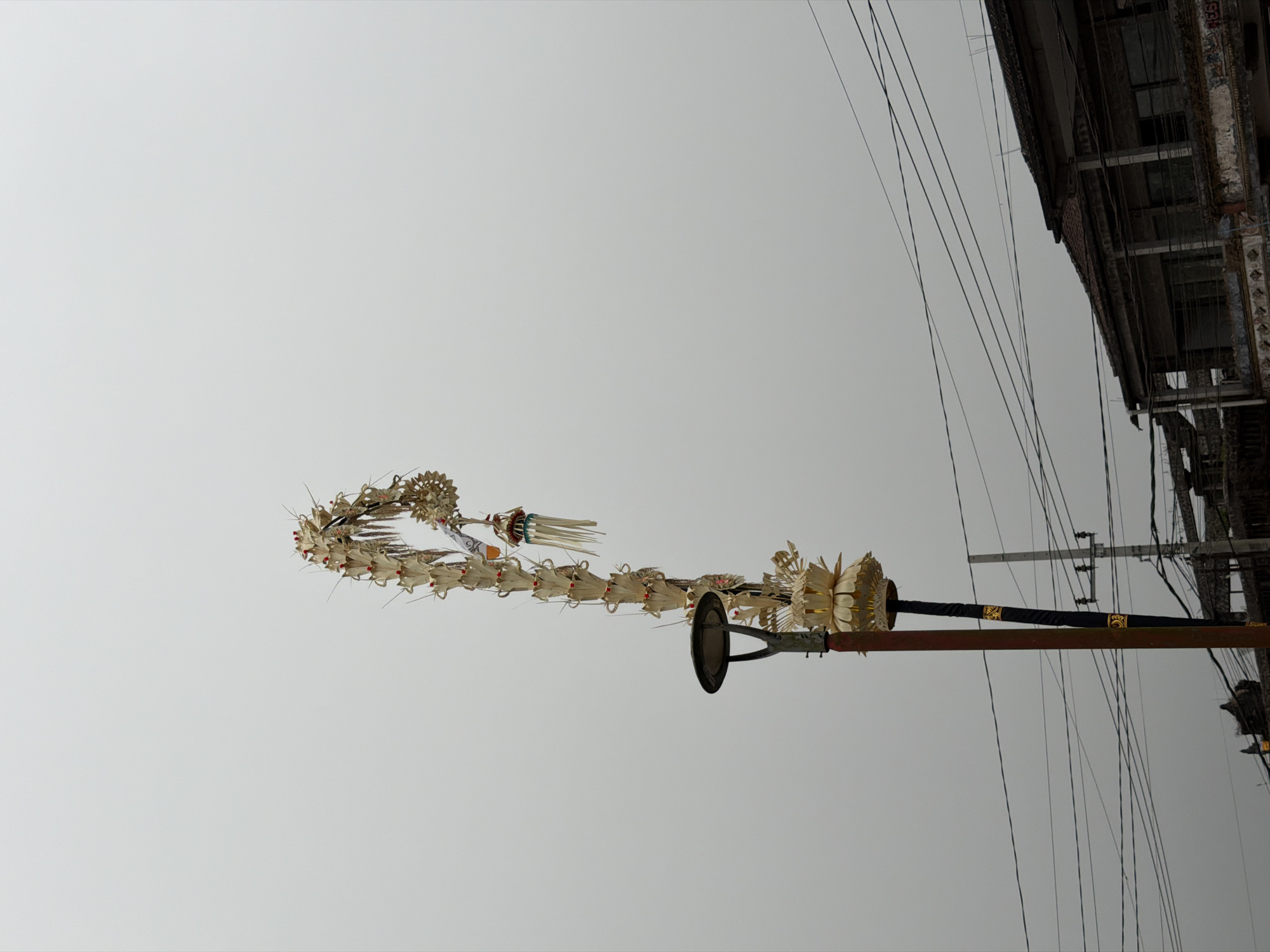 The image, captured in Kintamani, Indonesia, shows a tall, ornate pole adorned with numerous palm frond decorations, resembling a woven structure that curves upwards. At the base of this decorated pole, a street lamp fixture is visible. Below the decorated pole, a more utilitarian pole with black and gold banding is partially obscured. In the background, a two-story building with a tiled roof and weathered facade is visible. A network of power lines crisscrosses the sky above the building and the poles. The sky is overcast, creating a uniform, light gray backdrop.