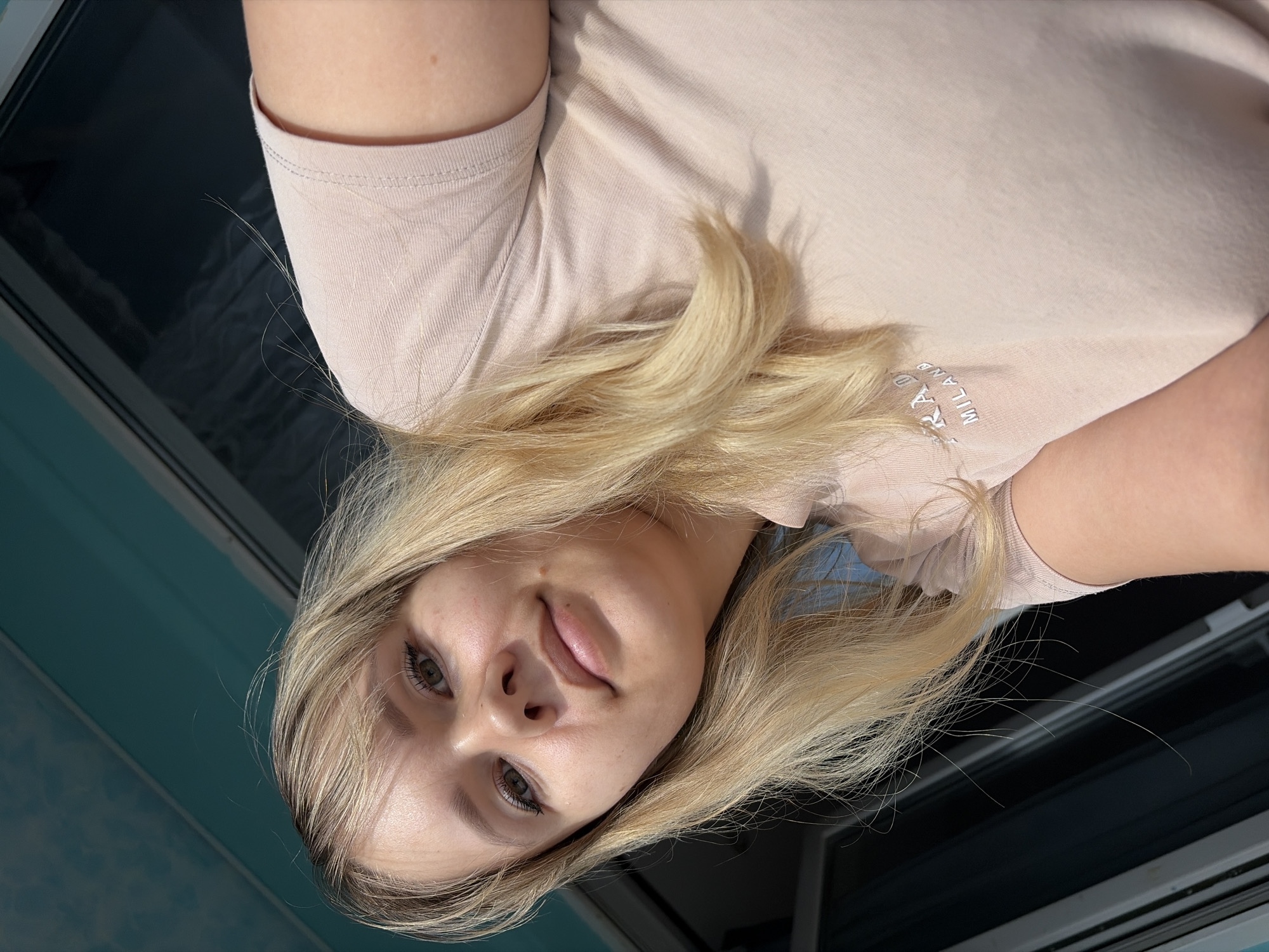 A young woman with blonde hair is seen in an indoor setting. She is wearing a light pink t-shirt with "PRADA MILANO" visible on the front. Her face is slightly tilted towards the camera. The background appears to be a room with an aqua-colored wall. The location is in the city of Unknown, Unknown.