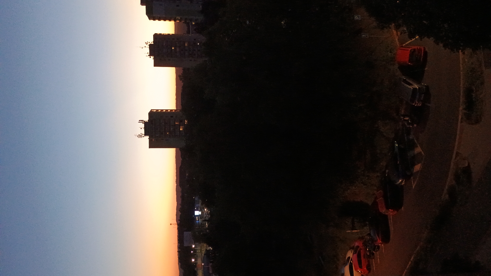 The image captures a twilight scene over the city of Sokolov, Czechia. The sky displays a gradient of colors, transitioning from a pale blue at the top to a warm orange and yellow horizon. Silhouetted against the sky are several tall apartment buildings, their windows faintly lit. In the foreground, dense, dark foliage obscures much of the ground level. A parking lot is visible at the bottom of the image, with several cars parked. The cars are various colors, including red, black, and silver. Some parking signs are also visible. The overall impression is a quiet evening in a residential area of Sokolov.