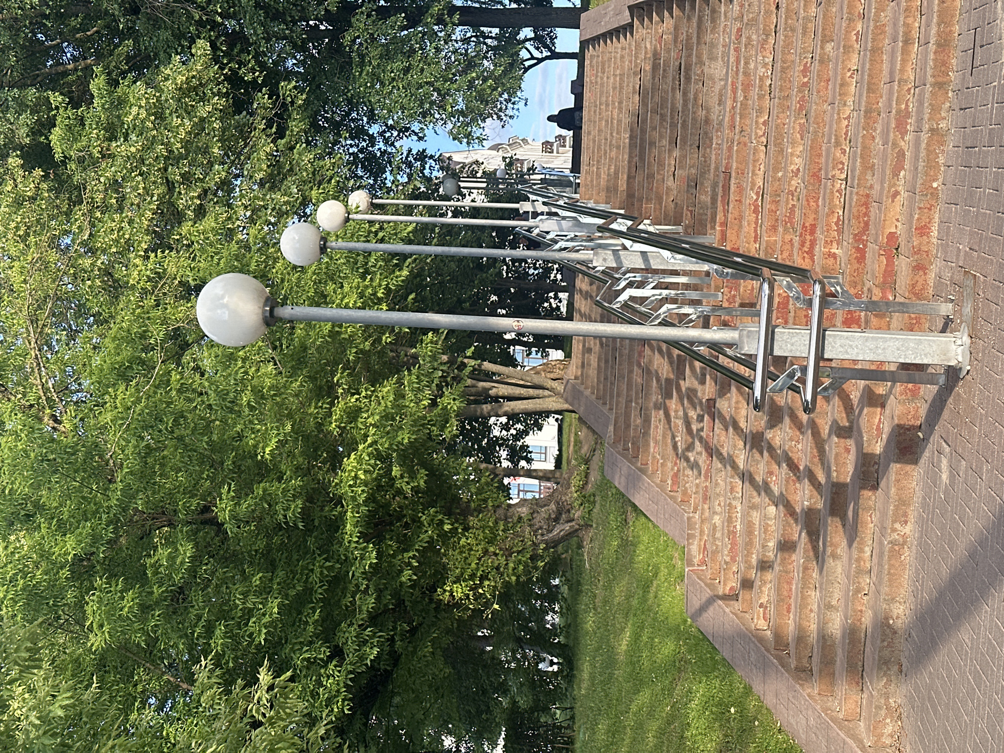 An outdoor stone staircase in Minsk, Belarus, ascends towards a building topped with a cupola. The steps are made of worn reddish-brown stone, with some areas showing chipped paint or material. A metallic handrail runs along the right side of the staircase, supported by silver posts. Several modern, spherical streetlights stand in a line along the left side of the stairs, casting shadows on the steps. Lush green trees with full foliage line the sides of the staircase and extend into the background, with dappled sunlight filtering through the leaves. A patch of green grass is visible to the left of the stairs. At the top of the stairs, a few people are visible near the building, with one person seated and another standing. The sky is a clear blue with a few wispy clouds. The ground at the bottom of the stairs is paved with brick-like tiles.