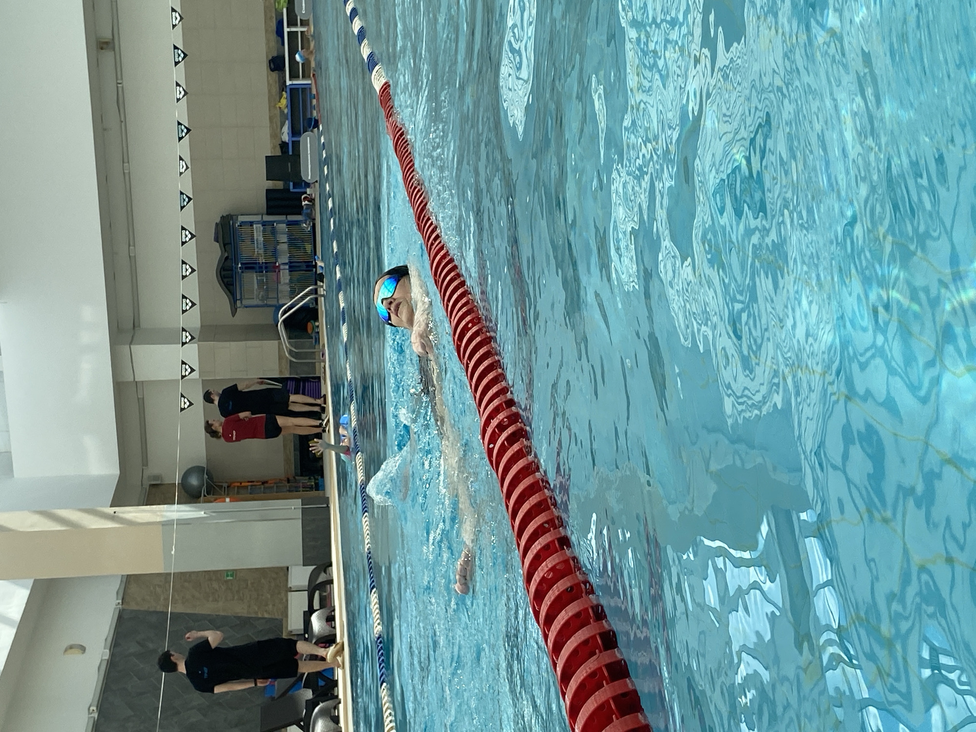 The image shows an indoor swimming pool. A person is swimming freestyle in a lane marked by a red and white floatation barrier. The swimmer is wearing goggles. There are other people standing at the edge of the pool. The pool appears to be in a modern facility with white walls and overhead lighting.
While the image shows an indoor swimming pool scene in Yekaterinburg, Russia, there is no definitive confirmation that the image was taken in Yekaterinburg.
