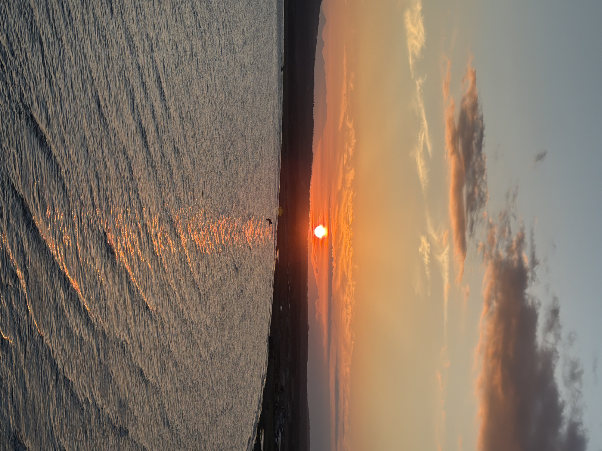 A high-angle, medium shot captures a sunset over the ocean near Yuzhno-Kurilsk, Russia. The sun, a bright orange orb, is partially obscured by thin, wispy clouds tinged with pink and orange, low on the horizon. The sky transitions from a pale blue at the top to a warm gradient of orange and yellow towards the horizon. In the distance, a dark, undulating treeline separates the sky from the water. The ocean's surface is calm, with gentle waves reflecting the sunset's warm glow, creating shimmering streaks of light. A single bird, silhouetted against the light, flies over the water. A few small structures are visible along the coastline, suggesting a populated area. The overall scene is serene and picturesque.