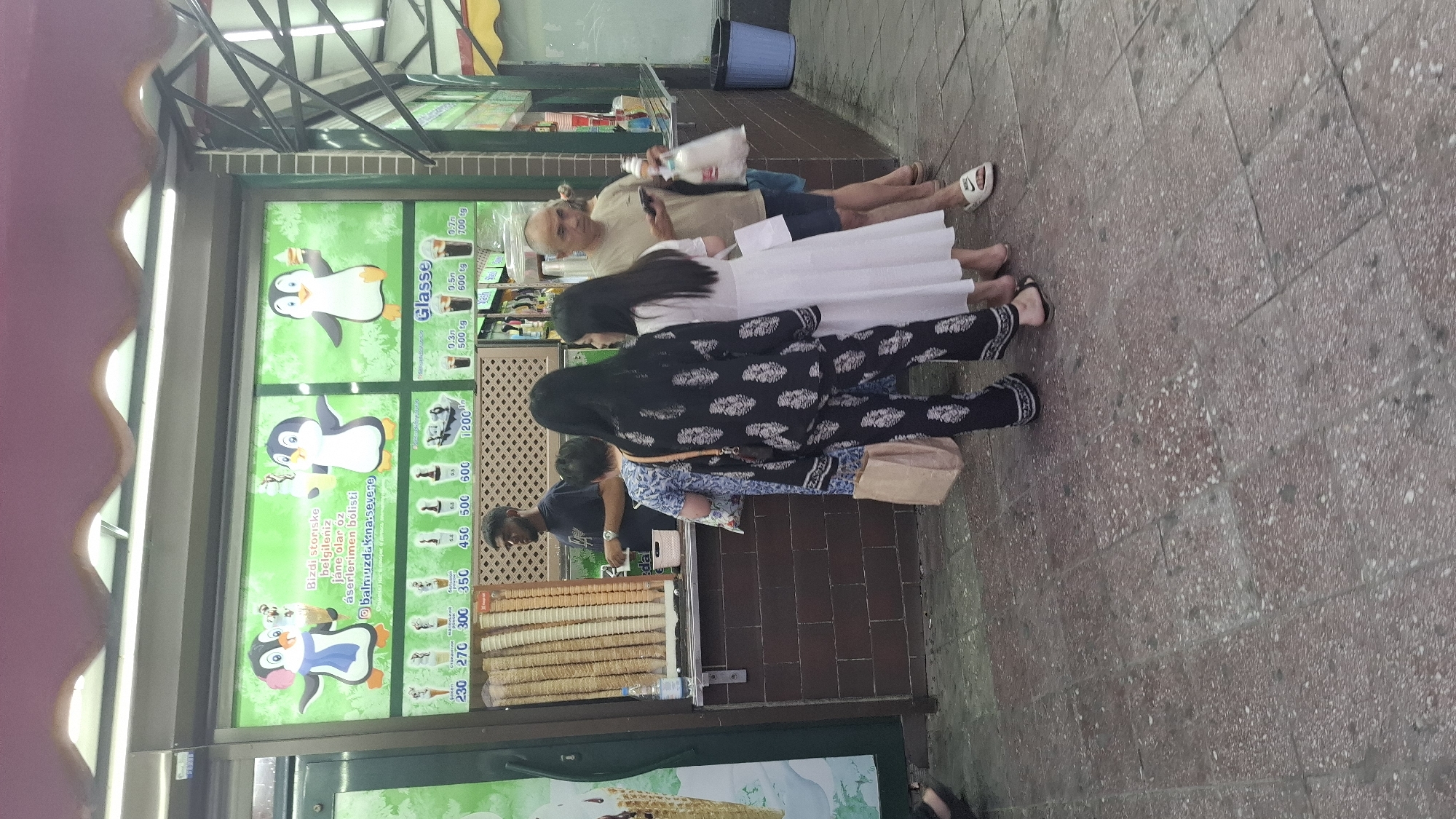 In Shymkent, Kazakhstan, an outdoor ice cream stand is depicted. The stand features a brightly lit menu board with images of penguins and various ice cream options. Below the menu, a large display of empty waffle cones is visible. A male employee, wearing a dark t-shirt, stands behind the counter, attending to customers. Three individuals are at the counter: a person in a blue and white patterned dress, and two individuals in dark patterned attire, one of whom is holding a bag. Further to the right, a man in a light-colored t-shirt and shorts is visible, holding a phone and a bag of ice cream. The ground is paved with reddish-brown tiles with a speckled pattern. The overall scene suggests a bustling commercial area on a warm day.