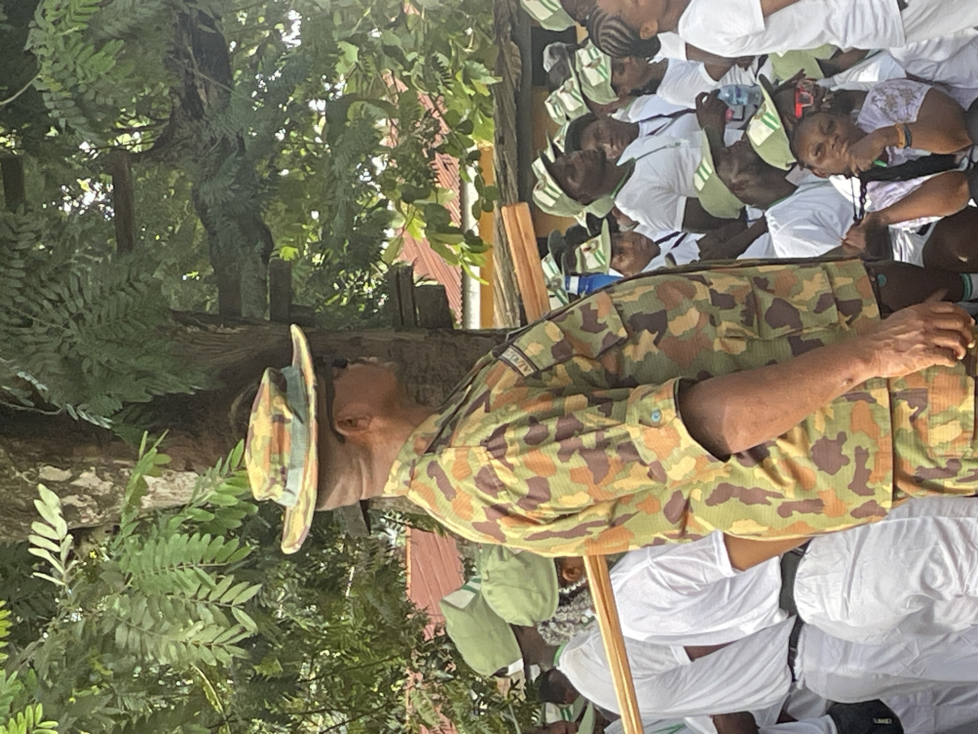 An outdoor scene in Ise, Nigeria, captures a gathering under bright daylight, likely midday or afternoon. In the foreground, a man in a camouflage military-style uniform and a matching boonie hat stands, his left side visible as he faces to the right of the frame. Partially visible text on his uniform sleeve appears to be "ADEN...".
Behind him, a group of young individuals, predominantly wearing white shirts and green and white caps, are seated. These caps, with their distinctive green and white color scheme and visible white logos, strongly suggest they are members of the National Youth Service Corps (NYSC), a mandatory one-year program for Nigerian graduates. The group appears attentive, with some individuals looking towards the front, possibly at the uniformed man. Some of the seated individuals also hold water bottles.
The setting is characterized by lush green foliage and trees, providing natural shade. In the mid-ground, through the trees, structures with red corrugated iron roofs are visible, typical of buildings in the region. A wooden stick or staff is partially visible in the lower foreground, near the uniformed man. The event seems to be a formal or semi-formal assembly, possibly an address or a briefing. The weather appears sunny and clear.