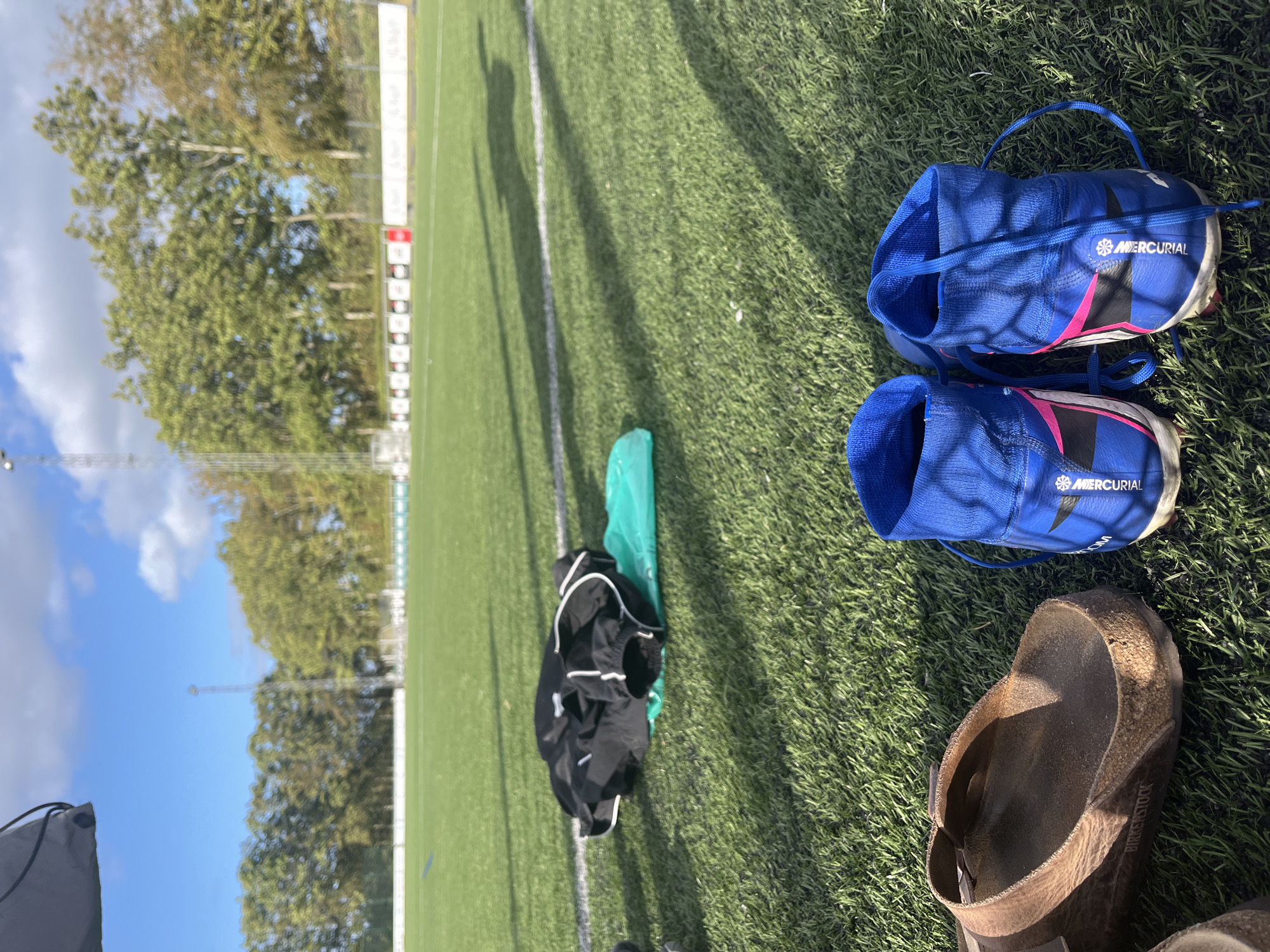 In Brejning, Denmark, on a sunny day, a pair of bright blue soccer cleats with white laces and pink accents rests on the artificial turf of a soccer field. To the left of the cleats, a brown leather Birkenstock sandal is visible. Further in the background, a black athletic bag lies on a bright green plastic bag on the grass. The field features white lines and is bordered by trees and a fence. A tall utility pole stands in the distance under a blue sky with scattered clouds.