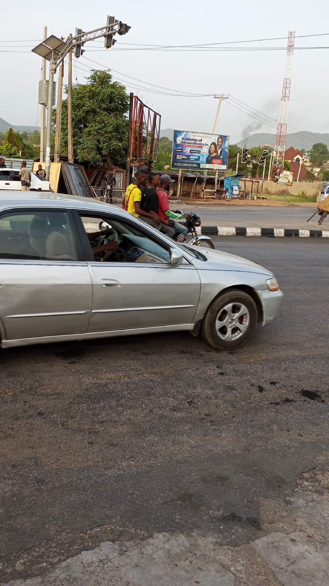 The image shows a street scene in Lokoja, Nigeria. A silver sedan occupies the left foreground, with a driver visible inside. To the right of the car, two men are riding a motorcycle. Further back, a group of men stands near a white vehicle. In the background, there is a large billboard, a telecommunications tower, traffic lights, and green hills. The road surface appears asphalt.