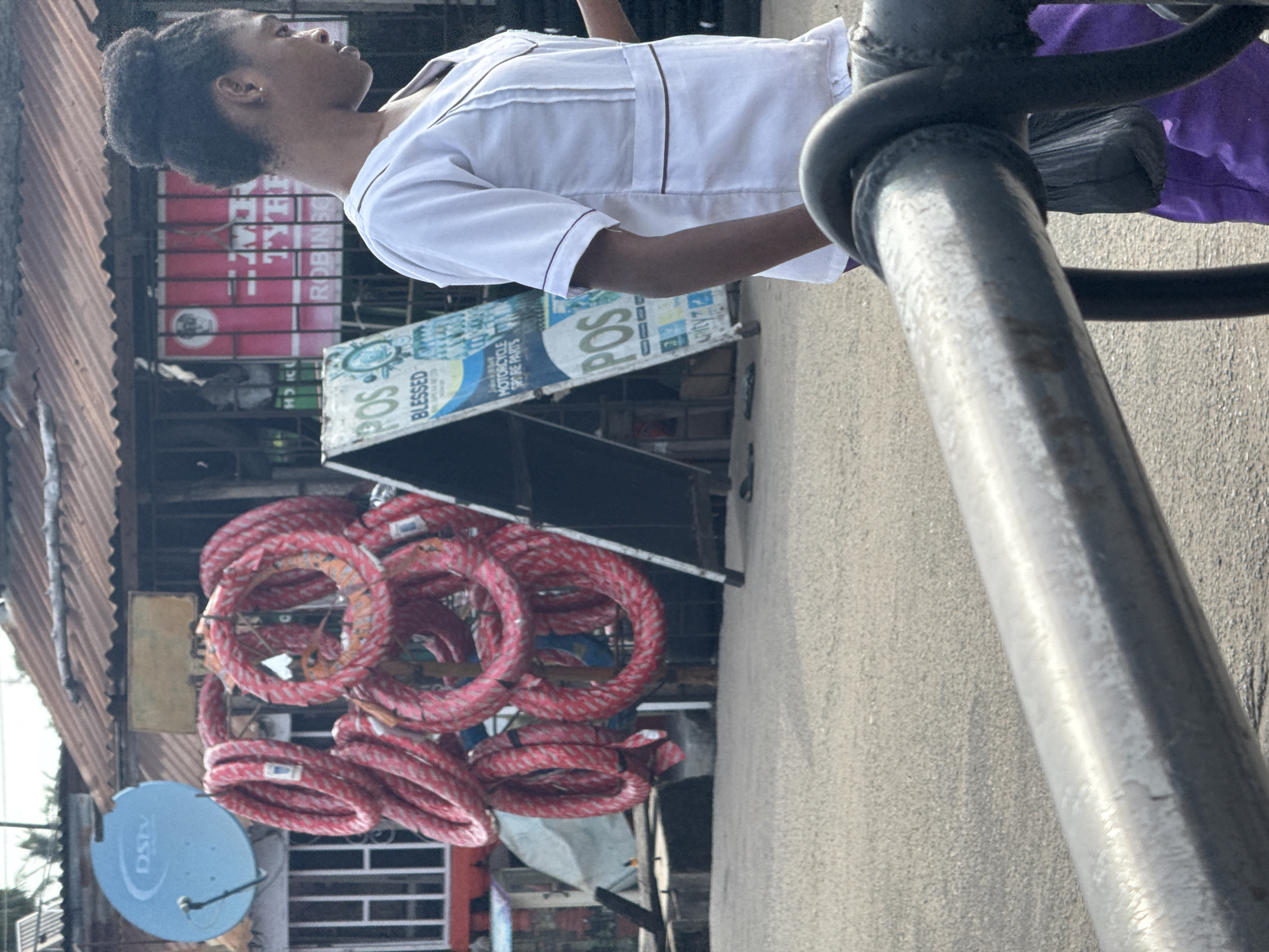 The image shows a young woman in Badagry, Nigeria. She wears a white, short-sleeved uniform with dark trim. She stands near a metal pipe structure on a street with signs, and a display of red, coiled materials. A blue Dstv satellite dish is mounted on a building on the left side of the image. The buildings appear to have corrugated iron roofs.
