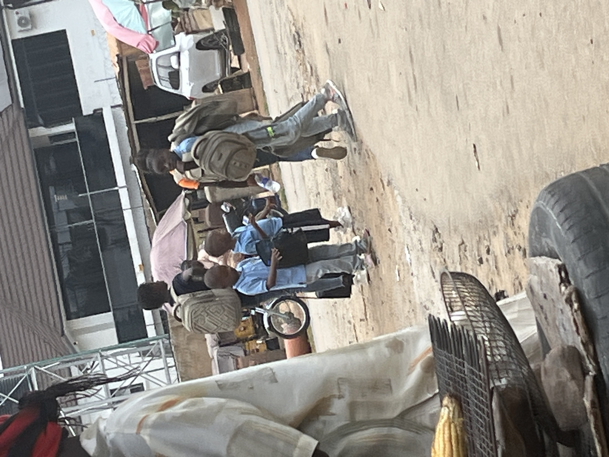 In a street scene in Abuja, Nigeria, a group of children, possibly students, are walking with backpacks. One child, wearing a light blue shirt and gray pants, carries a black bag. Another child, also in a blue shirt and gray pants, holds a backpack. To their right, a person in a dark shirt and gray pants walks with a large backpack slung over their shoulder, also carrying a black bag. In the foreground, a person in a white garment, partially visible from the side, stands near a grill with corn. The background reveals buildings with dark windows and a white SUV. The ground is dirt and gravel. The overall atmosphere suggests a bustling urban environment.