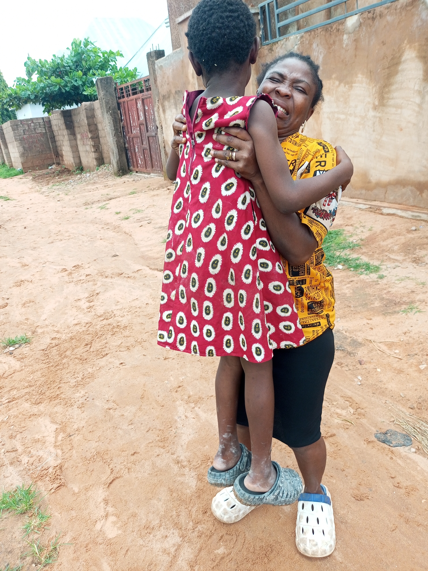 In Tempure, Nigeria, a Black woman is joyfully embracing a Black child. The woman, wearing a yellow shirt with intricate patterns and a black skirt, is seen with her eyes closed and a wide smile, her head tilted back as she hugs the child tightly. The child, dressed in a red dress with white and black circular patterns, is being held aloft by the woman, their back to the viewer. Both are wearing casual footwear; the woman has on what appears to be a black sandal, while the child is wearing a pair of gray clogs. The background shows a dusty, sandy outdoor area, with a partial view of a building with a pale yellow facade and a metal railing. To the left, a fence made of concrete blocks and a reddish-brown gate are visible, along with some lush green foliage. The ground is covered in fine, reddish-brown sand with scattered patches of dry grass and twigs. The overall scene conveys a moment of affectionate interaction between the two individuals in an outdoor, possibly residential, setting.