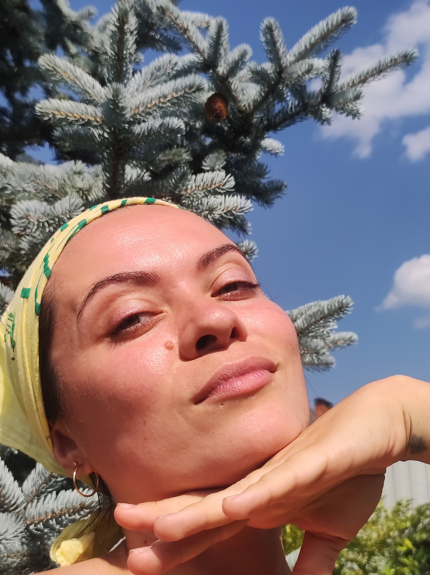 A close-up, low-angle selfie shows a woman with her head tilted back, resting her chin on her hand. She is wearing a yellow headscarf with green accents. She has a mole on her cheek and a small hoop earring in her left ear. Behind her, the blue sky is visible with a few white clouds. In the foreground and partially obscuring her face, is a branch of a blue spruce tree with a pine cone attached. The background appears to be a light-colored fence and some green foliage. The location is identified as Foki, Russia.