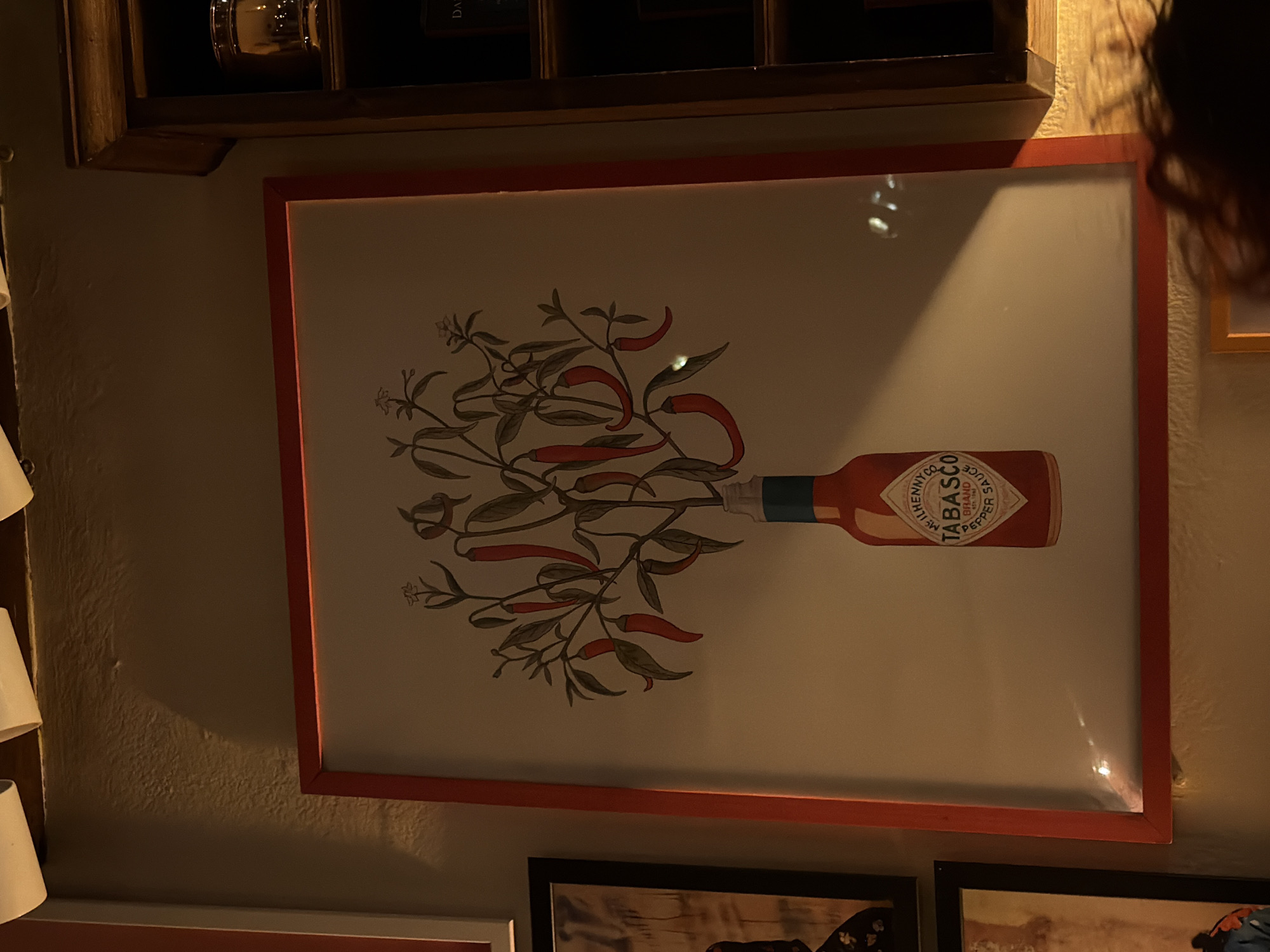 The image shows a framed print of a Tabasco sauce bottle with chili pepper plant features. It's hanging on a wall above other framed pictures, likely within an establishment. The location is Milan, Italy, although specific details tying it to that city are not readily evident in the picture. There is a person and some interior decor around the poster frame.