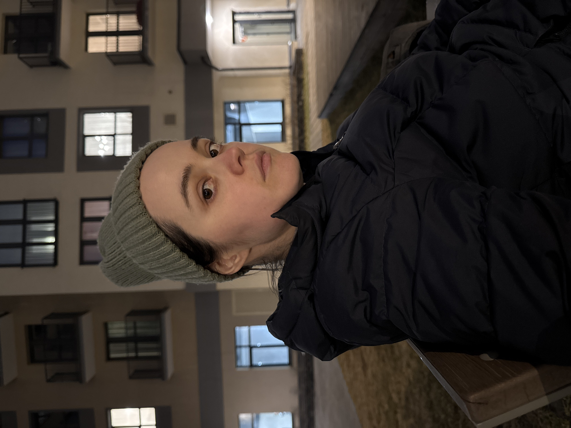 A woman wearing a green beanie and a black puffer jacket is sitting on a wooden bench outside. The background shows a multi-story building with illuminated windows. The photograph was taken in Izhevsk, Russia.