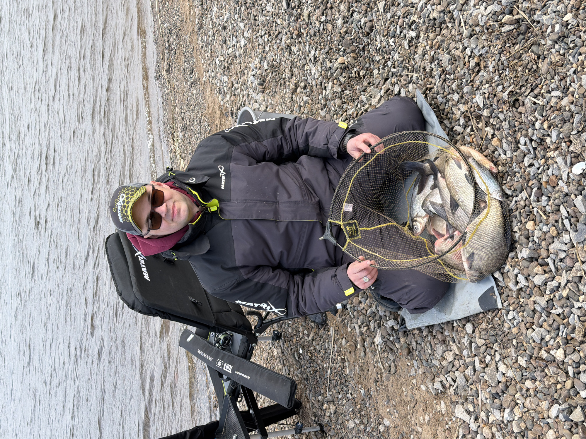 The image depicts a male individual sitting on a fishing chair by a pebbly shore after a successful fishing trip, set in Chaykovsky, Russia. The subject is seen from an elevated perspective, angled from the top-right.
He is dressed in a black, padded fishing jacket with distinctive yellow accents and multiple "MATRIX" brand logos. He wears dark sunglasses and a black and yellow patterned cap, also featuring the "MATRIX" logo. A silver ring is visible on his left hand.
The central focus of the image is a yellow-rimmed black fishing net, which the man is holding, filled with a substantial catch of freshwater fish, likely bream. The fish are laid out on a grey mat on the ground.
To the left of the fisherman, a black, sturdy fishing chair or seat box, branded "MATRIX," is set up on the rocky terrain. A fishing reel is visible on an attachment to the chair, and a black fishing rod bag or case, bearing partially visible text including "ULTRA FEEDER CONCEPT," "FOX," and "DURD," rests next to it.
The surrounding environment features a wide expanse of small grey and brown pebbles and rocks that constitute the shoreline. To the left, a large body of water, possibly a river or lake, appears calm with subtle ripples, reflecting a light grey tone under diffused lighting. The absence of harsh shadows suggests an overcast day or soft daylight. The scene captures the satisfaction of a good day's fishing.
