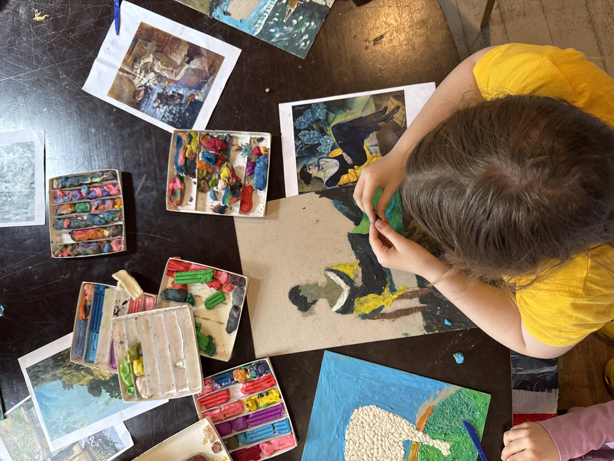 An overhead shot shows a child's hands working with modeling clay on a dark wooden table. Several opened boxes of colorful modeling clay are scattered around, along with printed images and partially completed clay art projects. One project features a textured white surface on a blue background. Another is a drawing of a figure in a black and yellow dress. The child, wearing a yellow shirt, is actively engaged in manipulating the clay. The scene appears to be a creative art session, possibly in a home or art studio setting. The location is not specified in the image or its context.