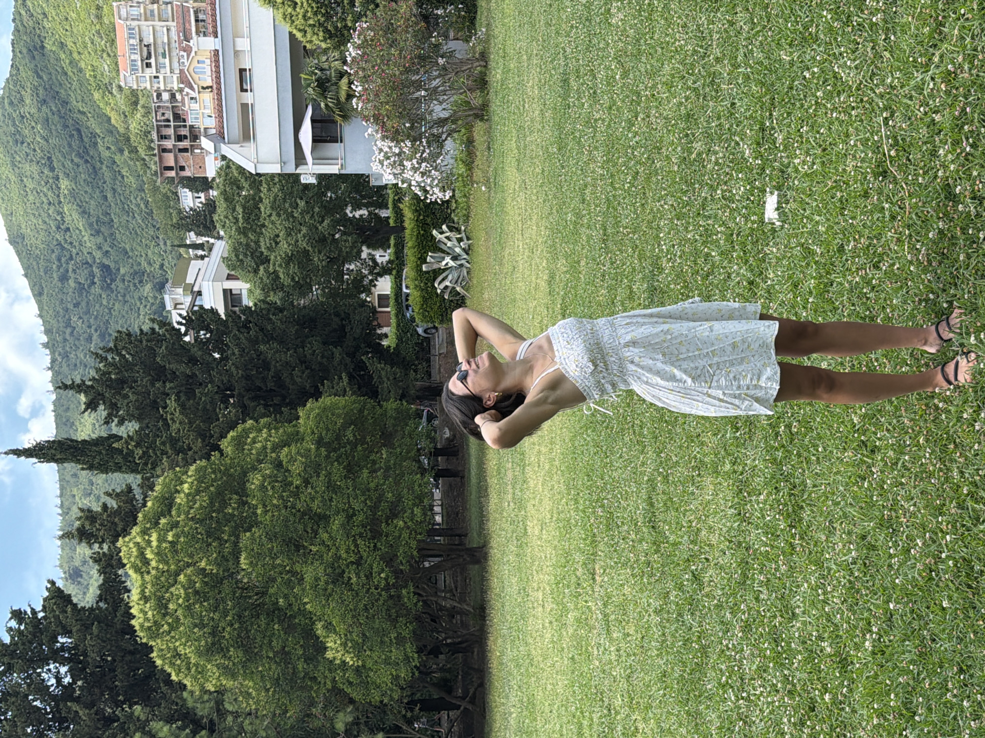 An outdoor, eye-level, full shot captures a young woman standing in a grassy area in Budva, Montenegro. She is facing slightly to her right, with her head tilted back and her hands clasped behind her neck, seemingly enjoying the warm weather. She is wearing a white, short, floral-patterned dress and dark sandals. Her dark hair is pulled back, and she has on dark sunglasses.
The background reveals a lush green landscape with various trees, including cypress and broadleaf varieties. In the middle ground, a few white flowering bushes add a touch of color. Beyond the trees, a hillside is dotted with buildings, some appearing to be under construction and others completed apartment buildings and houses, characteristic of a coastal town. The mountain slope behind the buildings is covered in dense green foliage. The sky above is partially clouded. The ground she stands on is covered in short, green grass interspersed with small white flowers, suggesting a well-maintained park or garden area. A white object, possibly a piece of paper or trash, is visible on the grass to her right.