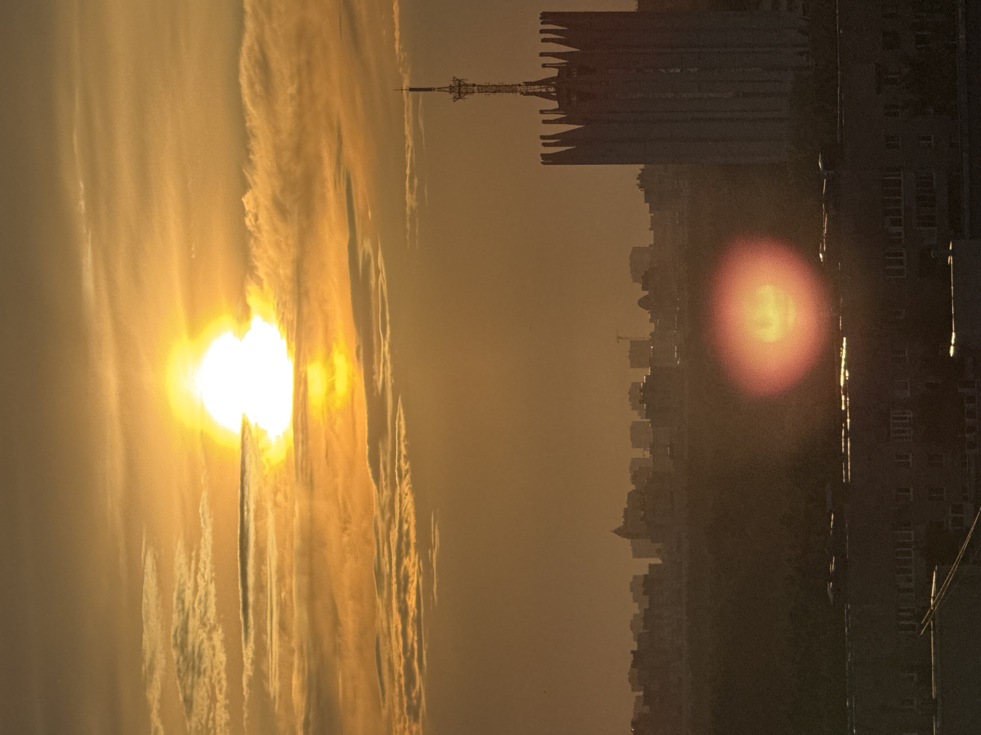 The image presents a dramatic sunrise or sunset over a sprawling urban landscape, with intense golden light dominating the scene. The sun, a brilliant orb of white and golden light, is prominently positioned on the left side, casting a warm, luminous glow across the sky. Streaky, elongated clouds, illuminated by the sun's fiery hues, extend across the upper and middle portions of the sky.
To the right, the city skyline appears as a dark silhouette against the bright backdrop. Numerous buildings create a jagged horizon line, and a distinct, tall, slender structure resembling a communication or TV tower rises above the other rooftops. In the immediate foreground on the far right, a large, dark building is partially visible, with faint patterns of windows catching some light. A notable reddish-orange lens flare is present in the mid-right section of the image, appearing as a soft, glowing circle.
The time of day is clearly either sunrise or sunset, indicated by the low angle of the sun and the golden light. The overall atmosphere is hazy, contributing to the diffused light and the striking contrast between the radiant sky and the shadowed city. There are no people visible, and no discernible text appears in the image. The location is an unknown city, consistent with the provided context of Russia.